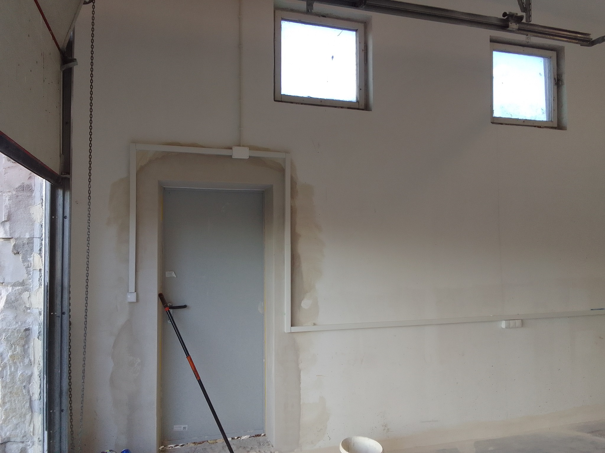 Wnętrze garażu w trakcie remontu: ściany przygotowane do malowania, widoczne ślady po szpachlowaniu i gładzi, okna w górnej części ściany, metalowa brama garażowa z łańcuchem, rura instalacyjna...