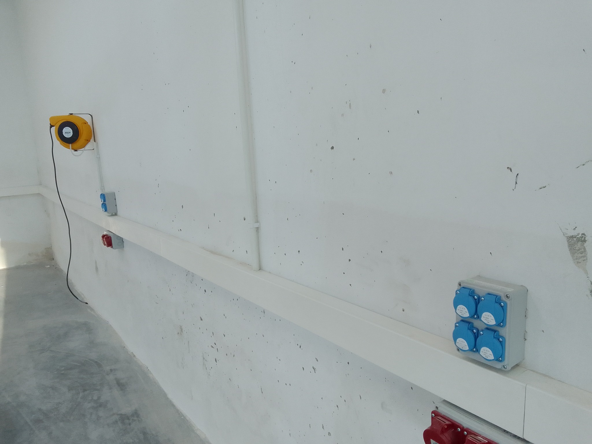Ściana z zamontowanymi gniazdami elektrycznymi przemysłowymi w kolorze niebieskim i czerwonym, umieszczonymi na białej listwie przypodłogowej. Dodatkowo widoczny zwijak kabla na ścianie.