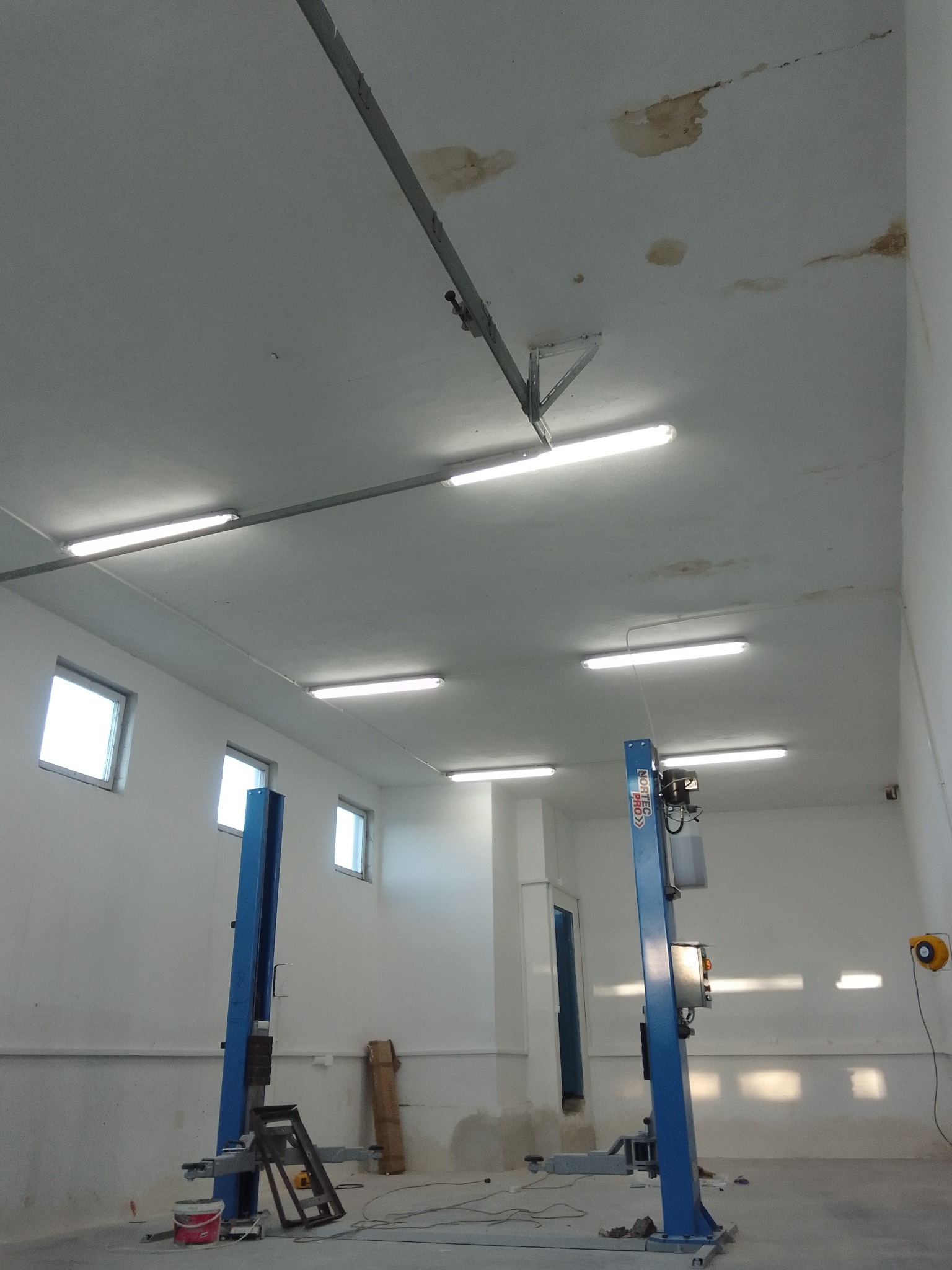 Wnętrze warsztatu samochodowego z dwoma niebieskimi podnośnikami, widoczne plamy na suficie i oświetlenie jarzeniowe.