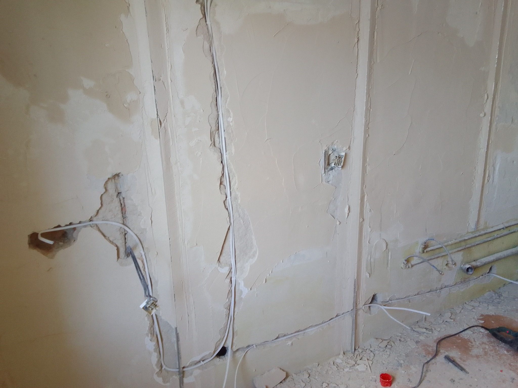 Ściana w trakcie remontu z widocznymi przewodami elektrycznymi wystającymi z otworów w ścianie, częściowo otynkowana, z widocznymi śladami po kuciu i resztkami gruzu na podłodze.