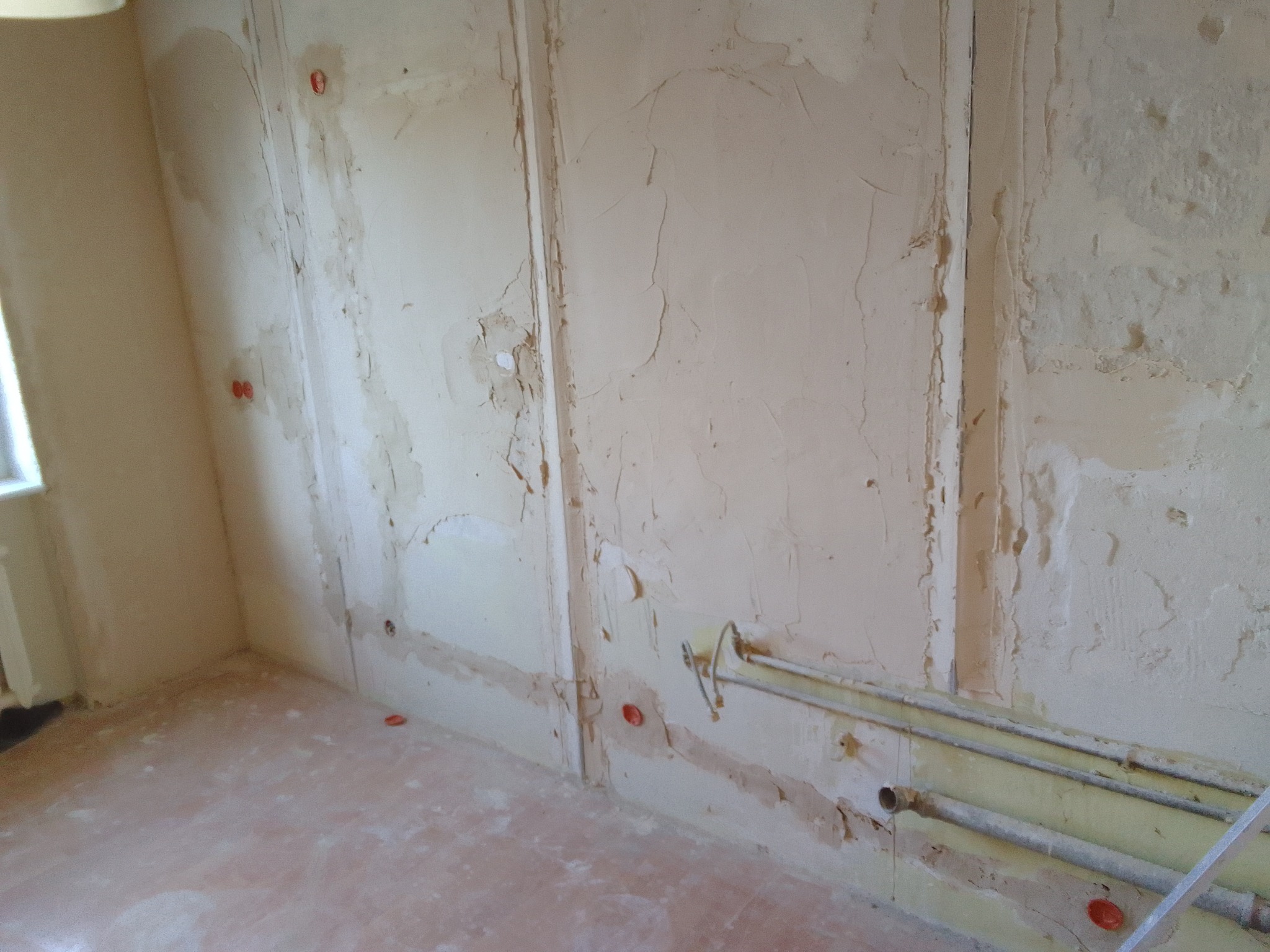 Pokój w trakcie remontu: ściany z widocznymi śladami po instalacjach, częściowo pokryte gładzią, rury grzewcze zamontowane wzdłuż ściany, podłoga z resztkami materiałów budowlanych.