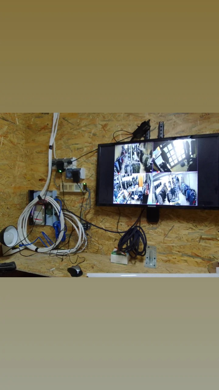 Ekran monitora prezentuje widok z kamer monitoringu w sklepie odzieżowym, obok widoczne kable, zasilacze i lampa na ścianie z płyty OSB.