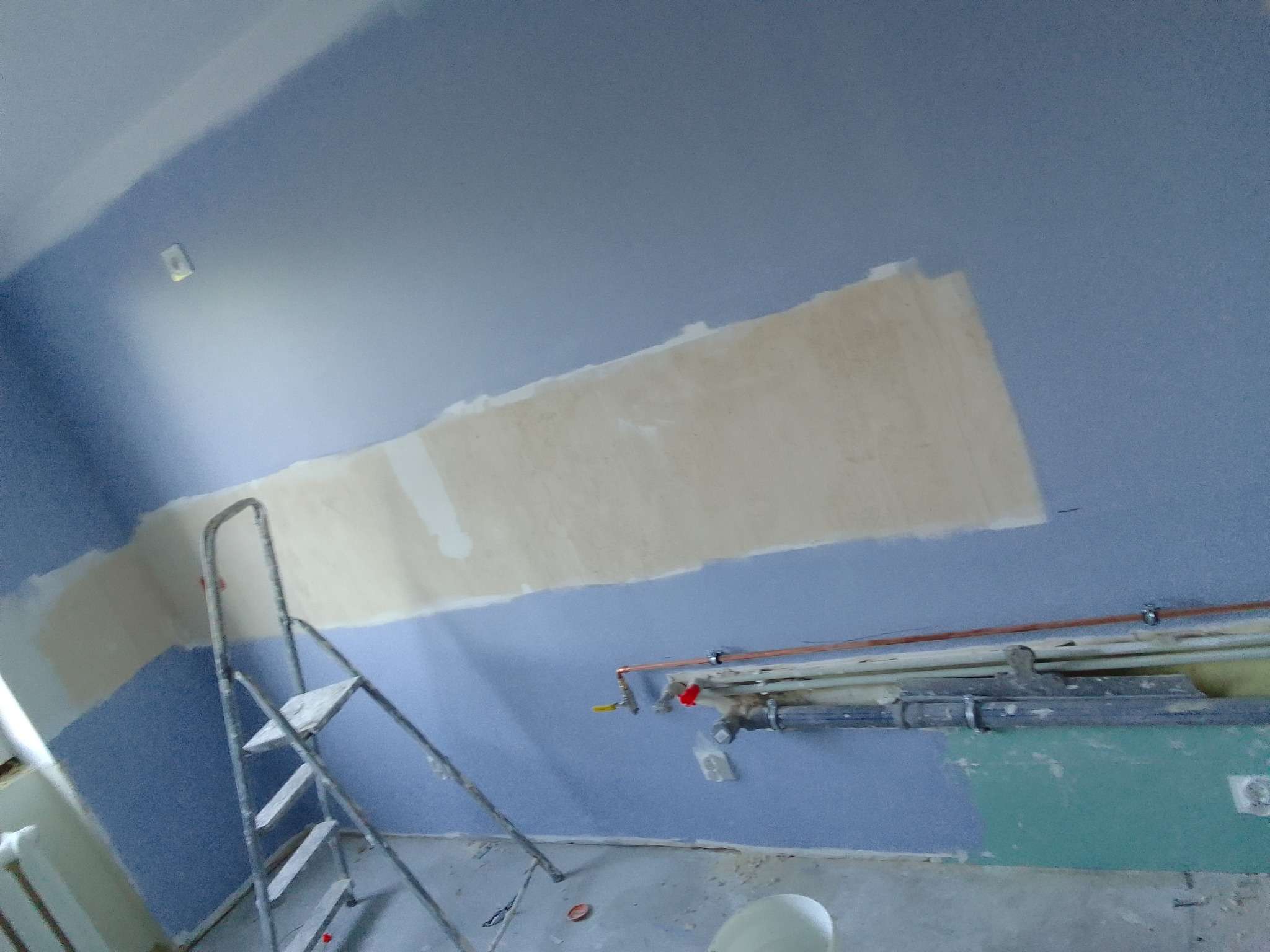 Niedokończona ściana pomalowana na niebiesko z fragmentem zakrytym płytą kartonowo-gipsową, widoczna drabina malarska i rury wzdłuż ściany.