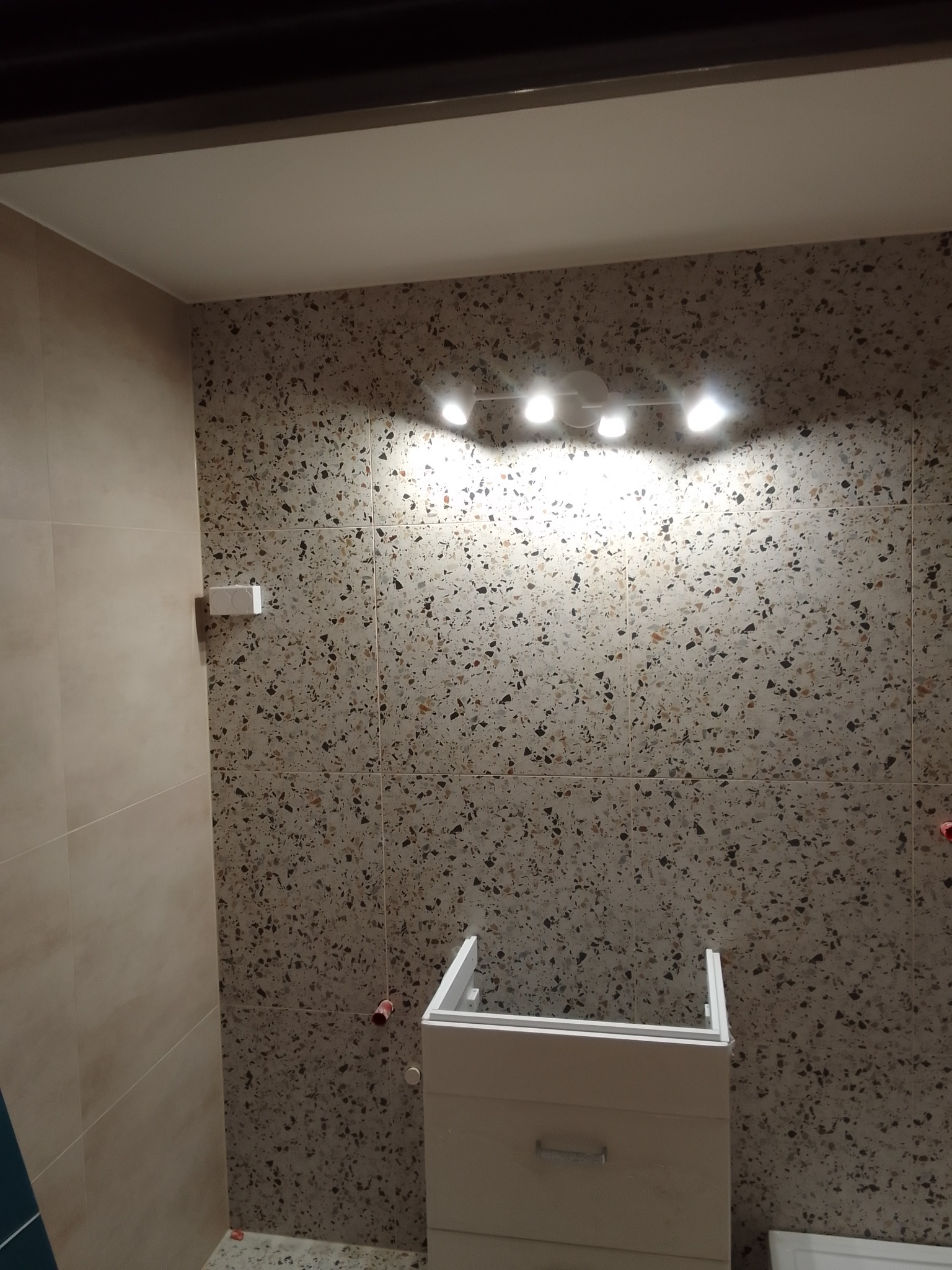 Łazienka w trakcie wykańczania: ściany wyłożone płytkami z motywem lastryko, zamontowane oświetlenie punktowe, częściowo zamontowana szafka pod umywalkę, widoczne wystające rurki instalacji wodnej.