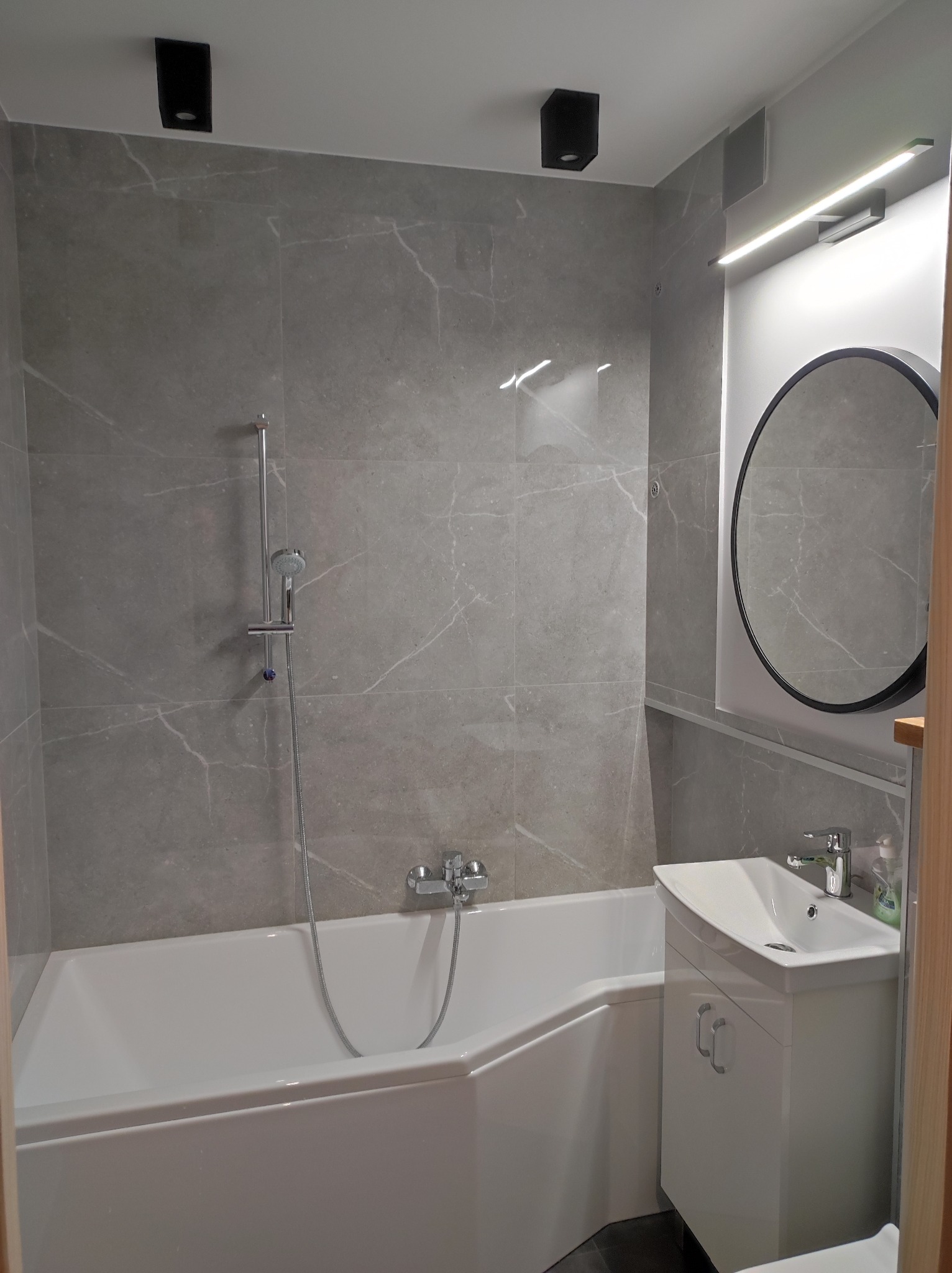 Nowoczesna łazienka z wanną obudowaną płytkami imitującymi marmur, białą szafką pod umywalką i okrągłym lustrem podświetlonym od góry.