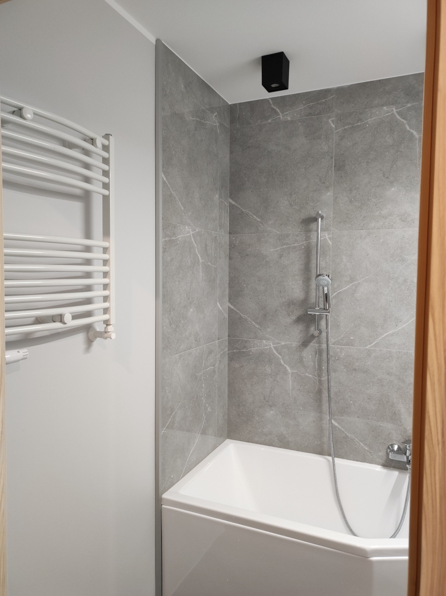 Wyremontowana łazienka z wanną obłożoną szarą marmurową płytką, chromowaną baterią i deszczownicą, biały grzejnik drabinkowy na ścianie, czarny punktowy reflektor sufitowy.