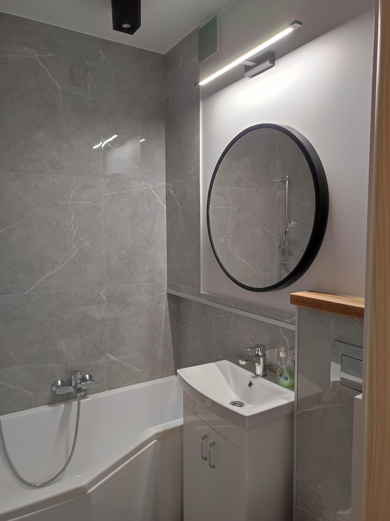 Nowoczesna łazienka z wanną, umywalką z szafką, okrągłym lustrem i szarą marmurową okładziną ścienną.