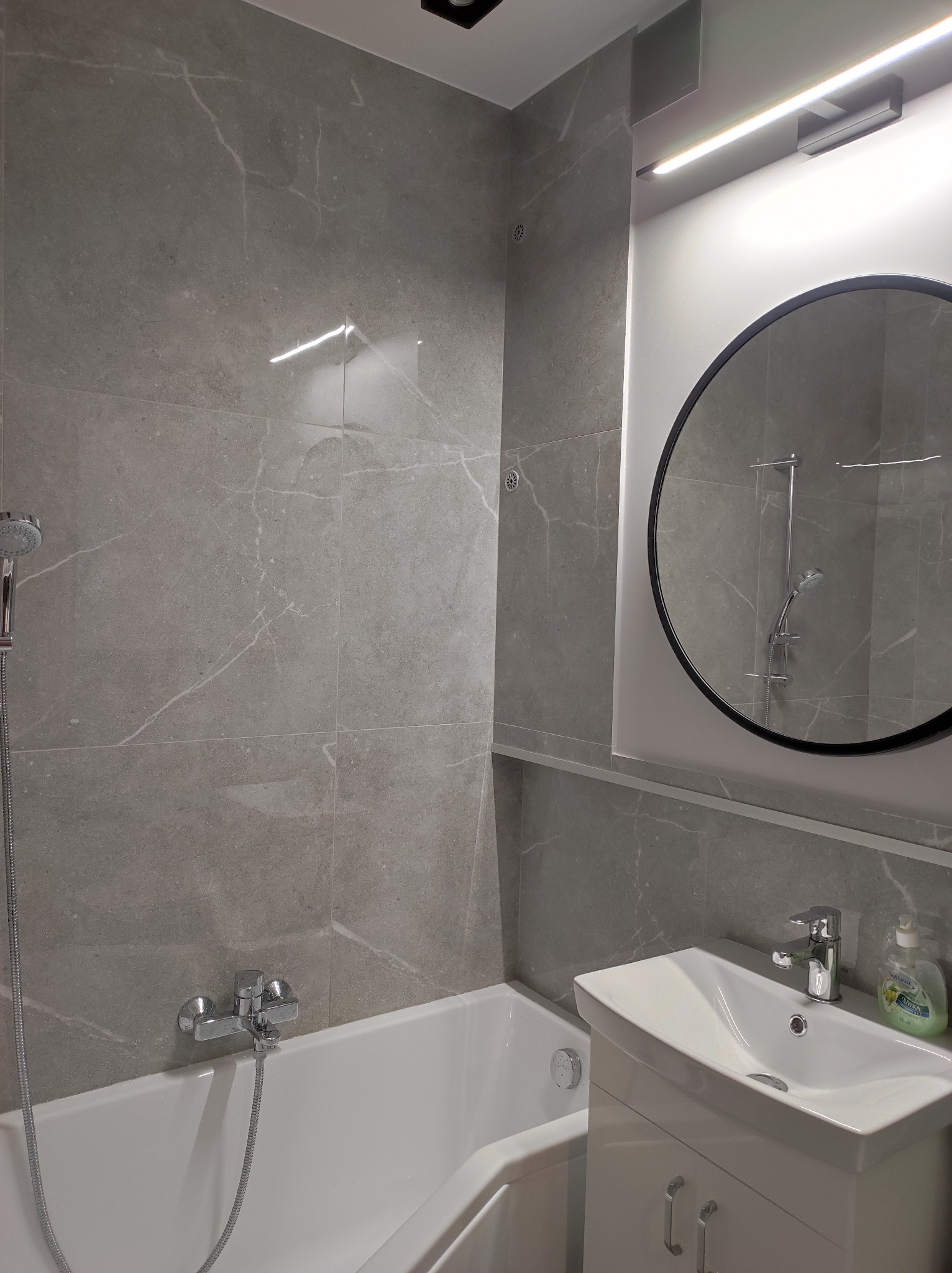 Nowoczesna łazienka wykończona szarymi płytkami imitującymi marmur, z owalnym lustrem w czarnej ramie, białą umywalką z szafką i wanną z chromowaną armaturą.