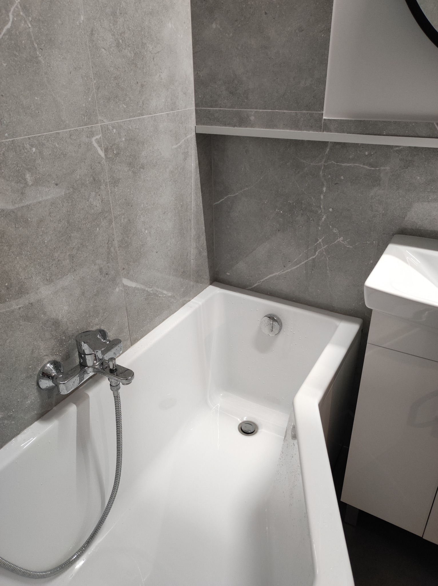 Nowoczesna, wykończona łazienka z wanną, szarą marmurową okładziną ścienną i białą szafką pod umywalką.