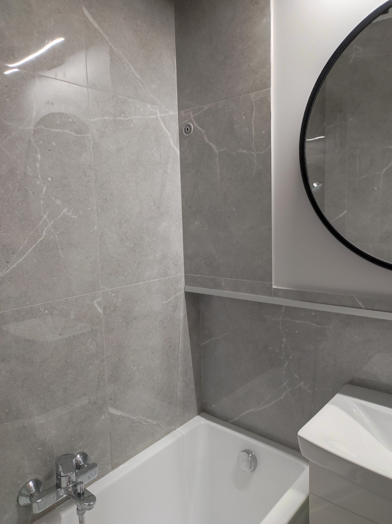 Wykończona łazienka z szarymi płytkami imitującymi marmur, okrągłym lustrem z czarną ramą nad umywalką i białą wanną z chromowaną baterią.
