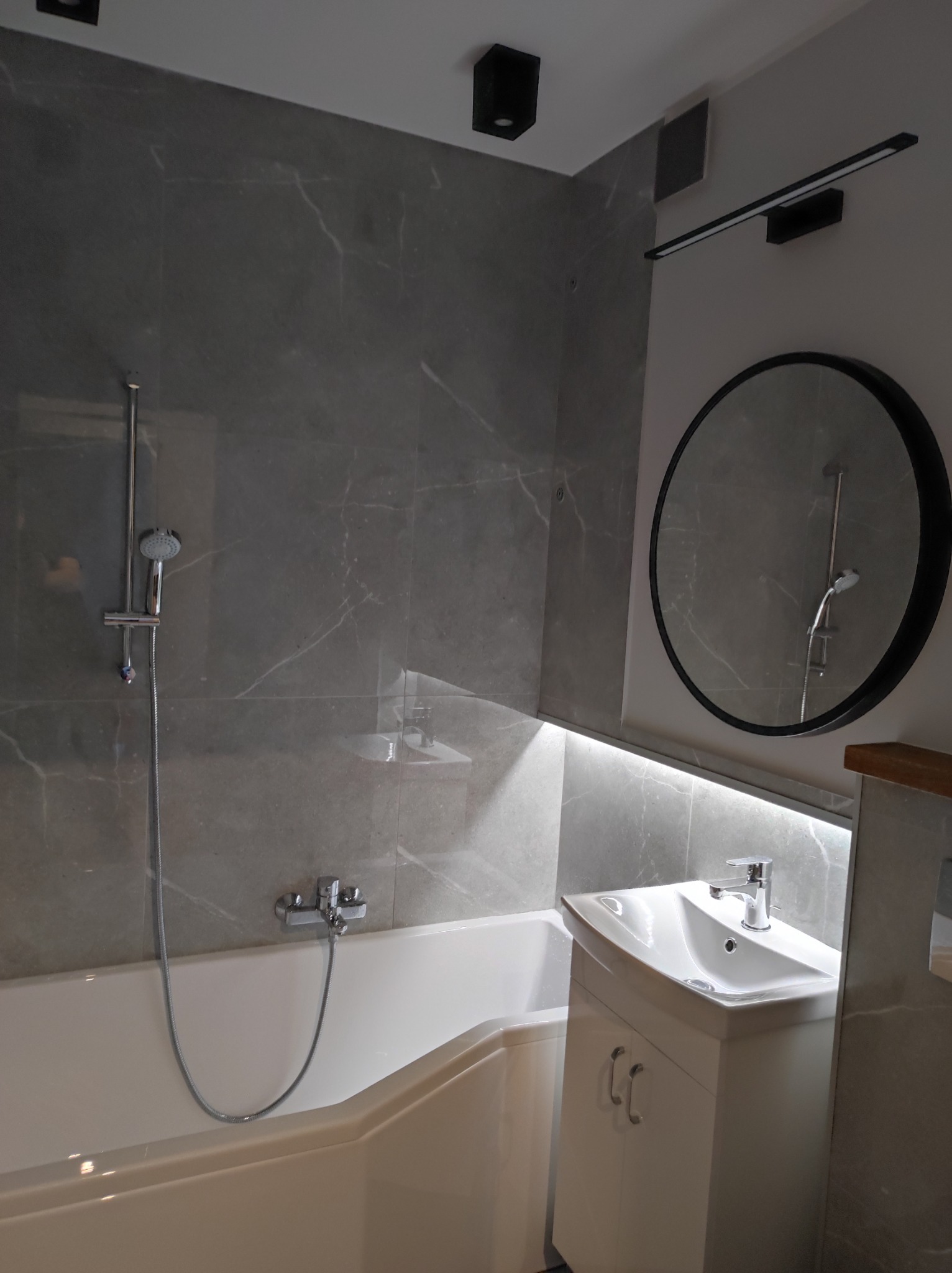 Nowoczesna łazienka z wanną, umywalką z szafką, okrągłym lustrem i szarymi płytkami imitującymi marmur, oświetlona punktowymi lampami sufitowymi i listwą LED nad umywalką.