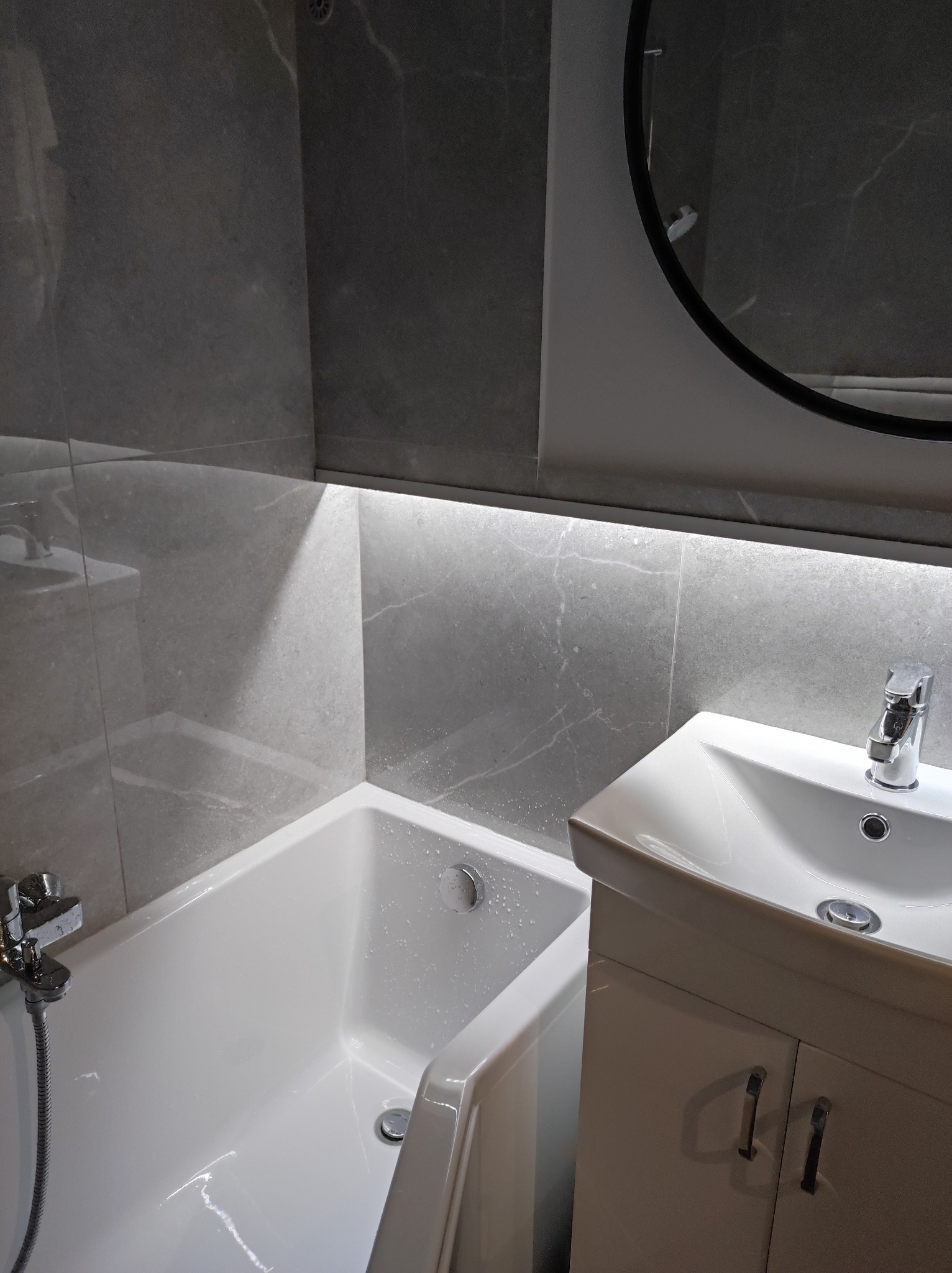 Nowoczesna łazienka z wanną, umywalką z szafką i podświetlanym lustrem w czarnej ramie, wykończona szarymi płytkami imitującymi marmur.