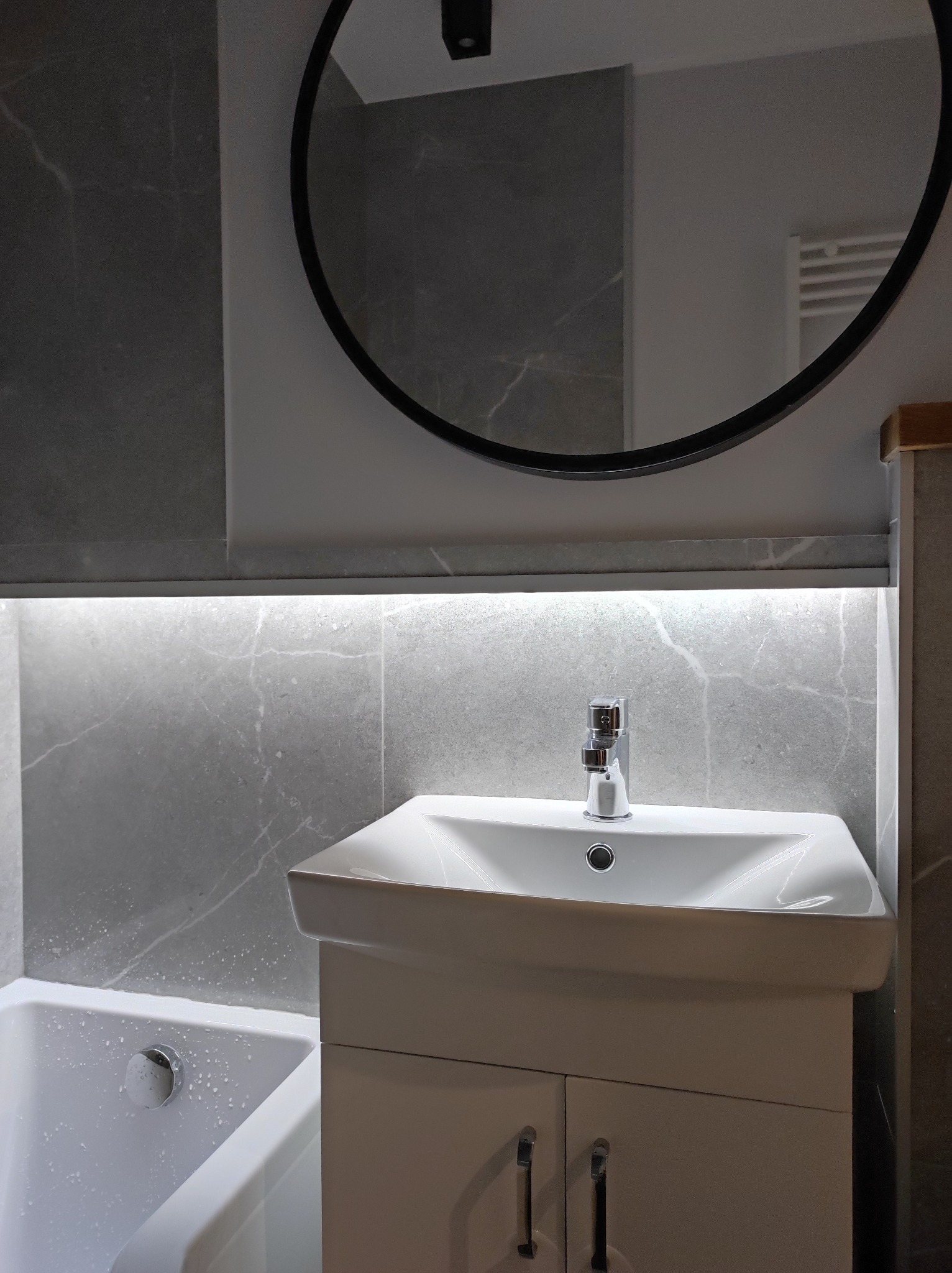 Nowoczesna łazienka z umywalką na szafce, bateria chromowana, okrągłe lustro z czarną ramą i podświetleniem LED nad umywalką, szare płytki imitujące marmur na ścianach i fragment wanny.