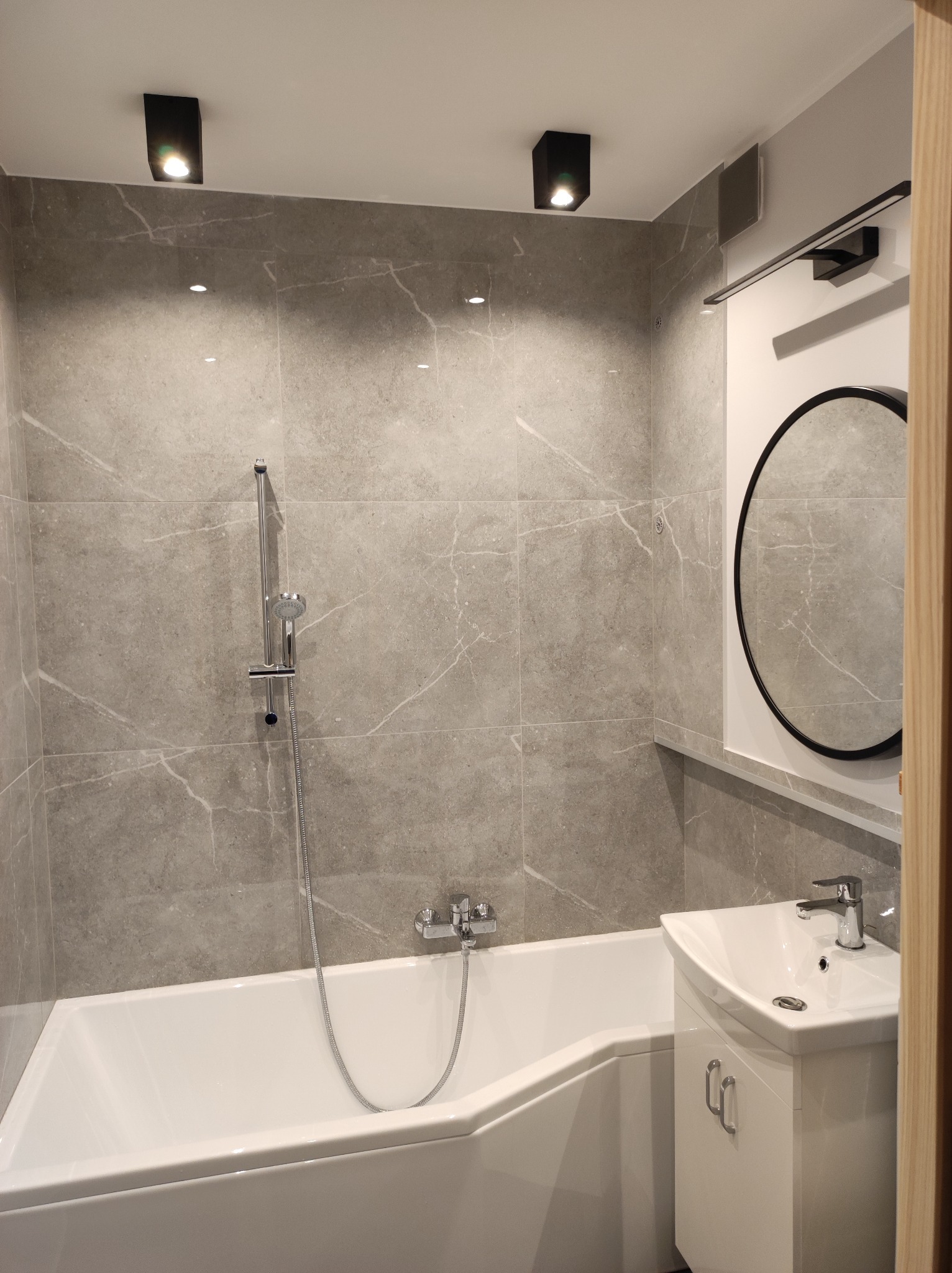 Nowoczesna łazienka w Łodzi z wanną, umywalką z szafką i szarymi płytkami imitującymi marmur, oświetlona punktowymi lampami sufitowymi oraz podłużnym oświetleniem nad okrągłym lustrem.