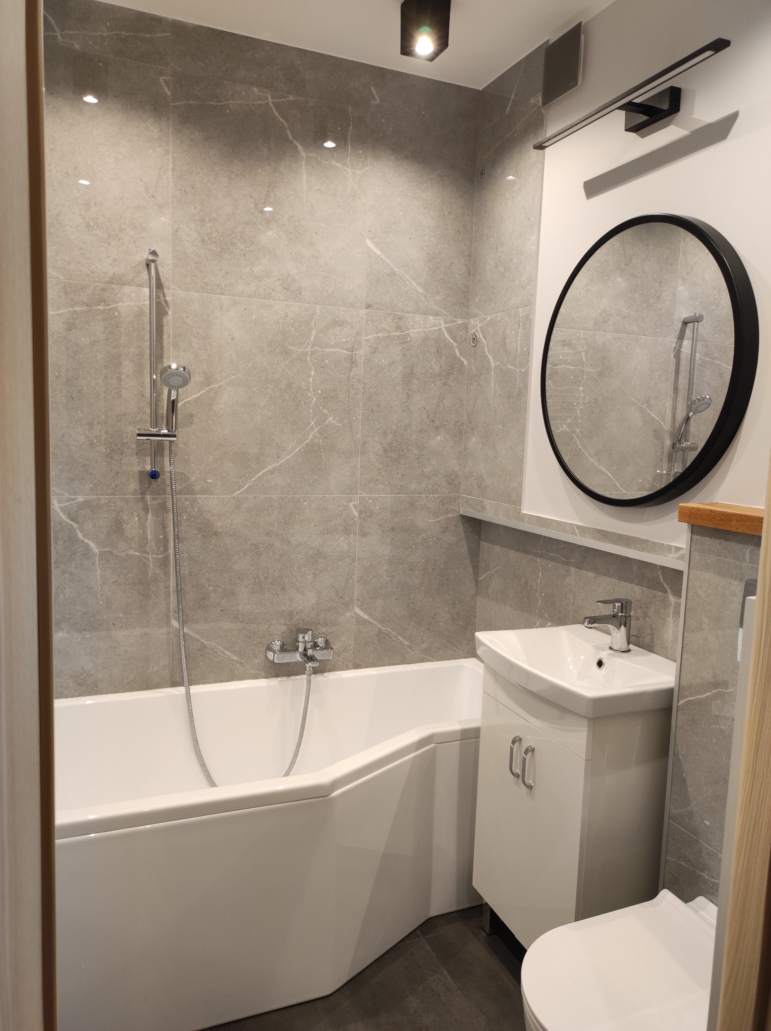 Nowoczesna łazienka z wanną narożną, umywalką z szafką, okrągłym lustrem w czarnej ramie i szarą marmurkową glazurą na ścianach.