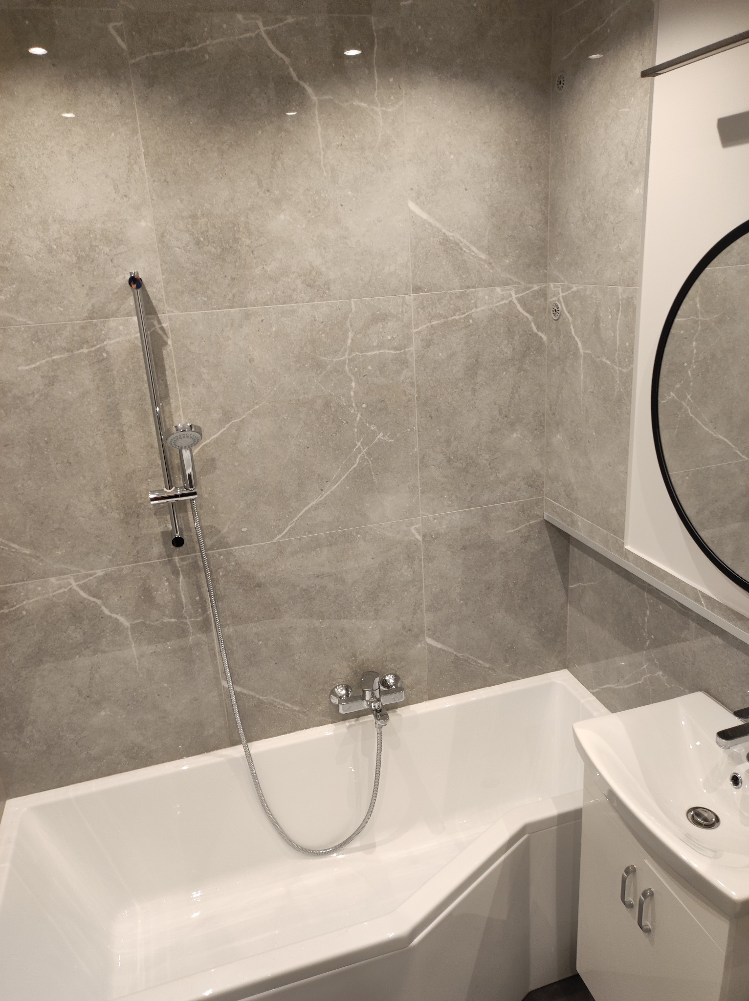 Nowoczesna łazienka z wanną narożną, umywalką z szafką i szarymi płytkami imitującymi marmur, oświetlenie punktowe w suficie.