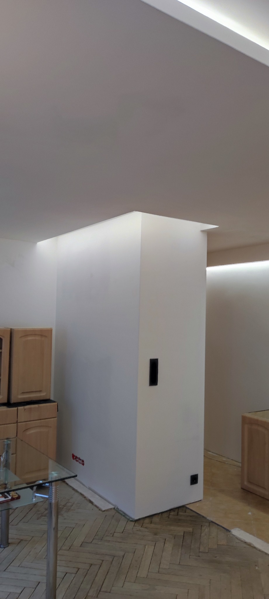 Nowoczesne oświetlenie sufitowe LED w odnowionym wnętrzu z drewnianą podłogą w jodełkę, widoczne meble kuchenne i fragment szklanego stołu.