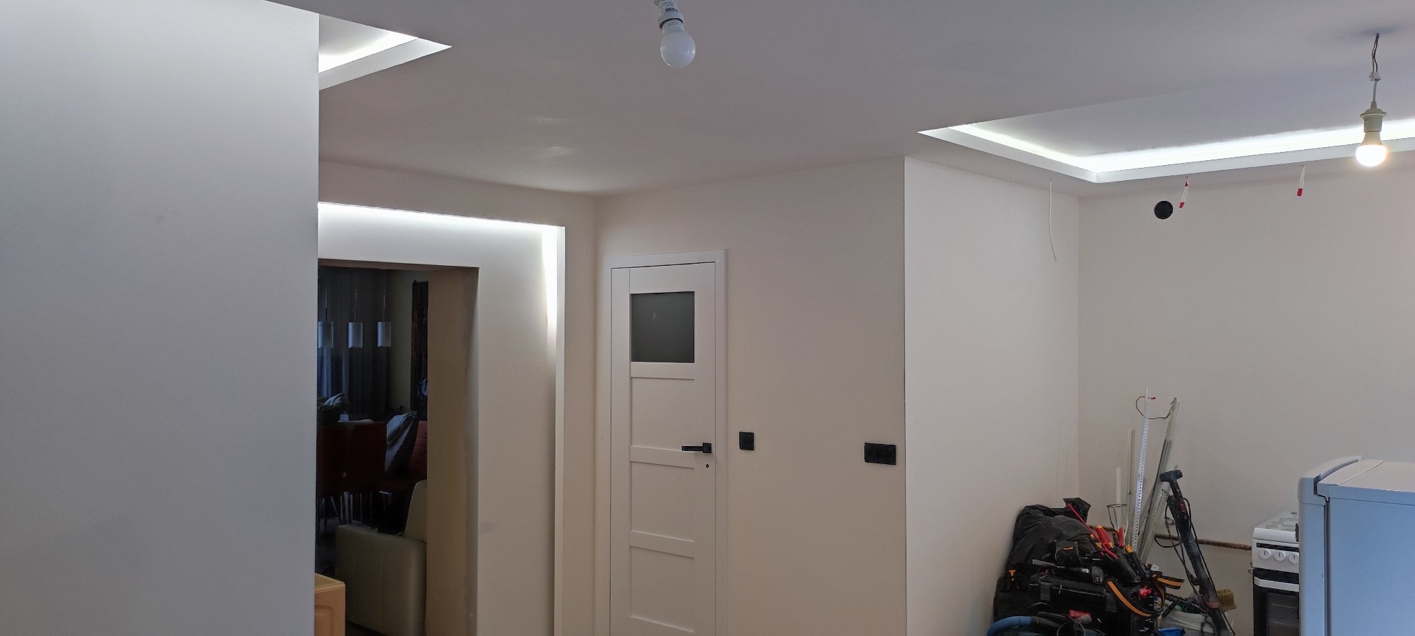 Wykończone wnętrze po remoncie: białe ściany, sufit z oświetleniem LED, białe drzwi z szybą, widoczne gniazdka elektryczne, w tle częściowo widoczne meble i sprzęt budowlany.