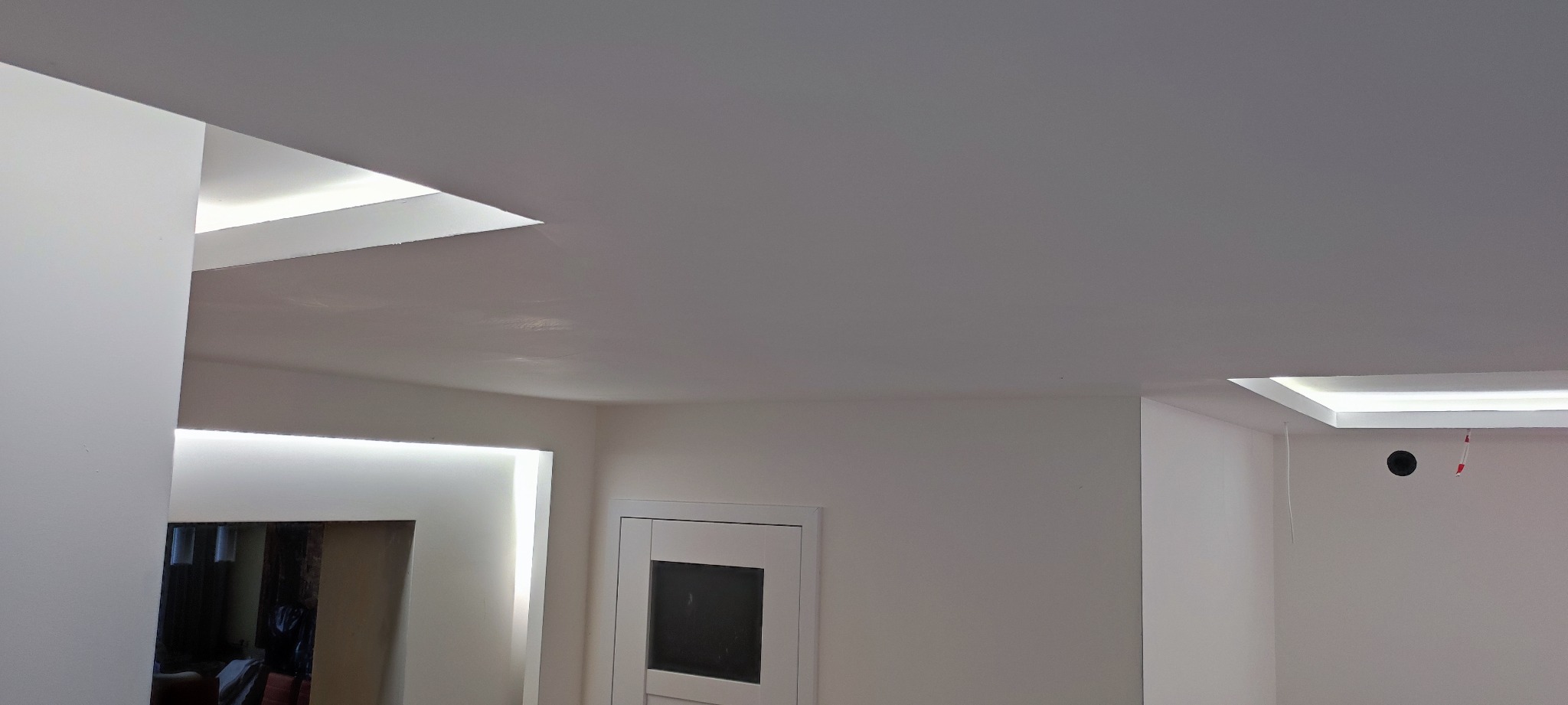 Minimalistyczne wnętrze: białe ściany, szary sufit z wbudowanym oświetleniem LED w geometrycznych wnękach, widoczny fragment drzwi.