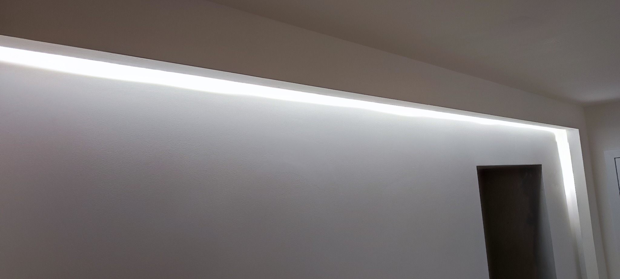 Biała ściana z wnęką oświetloną listwą LED, widoczny fragment sufitu i framuga drzwi.