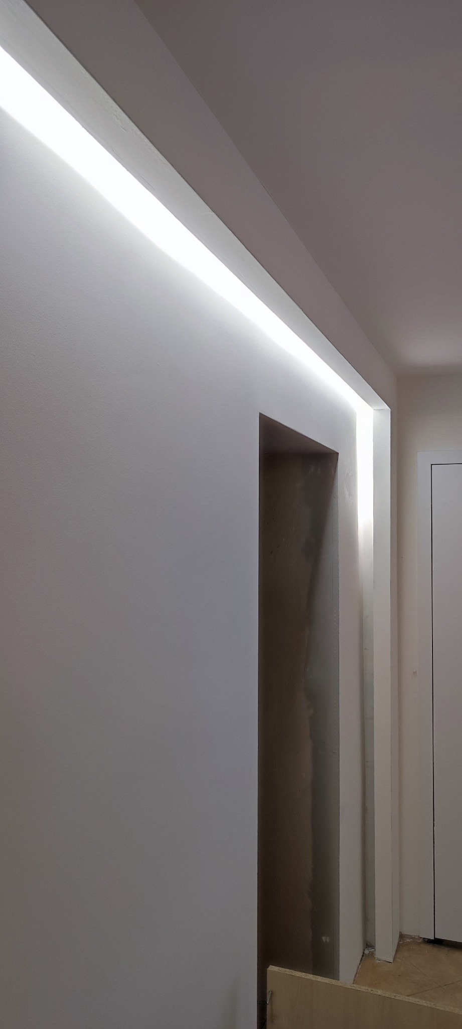 Biała ściana z oświetleniem LED wzdłuż sufitu, widoczne surowe wykończenie wokół otworu drzwiowego, częściowo wyszpachlowana ściana.