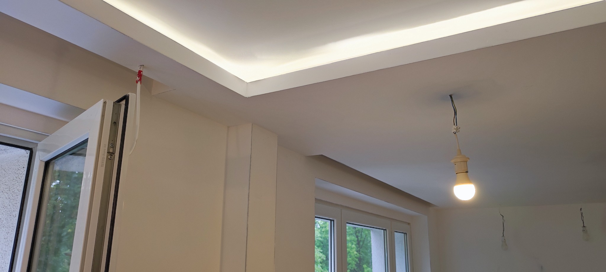 Nowoczesny sufit z oświetleniem LED w listwie, widoczne kable i prowizoryczne oświetlenie żarówką, okno w tle.