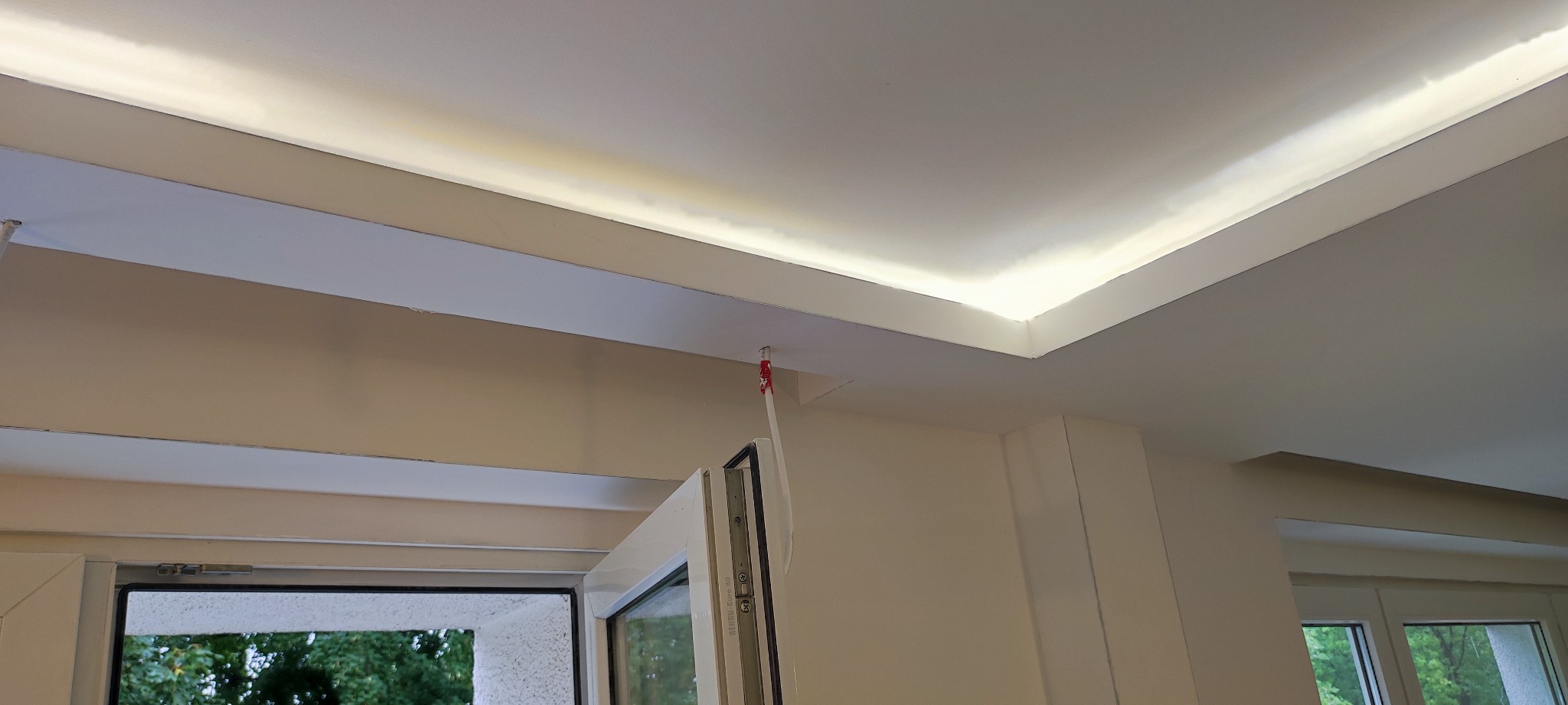 Nowoczesny sufit podwieszany z wbudowanym oświetleniem LED w jasnym pomieszczeniu, widoczne okno z zielenią na zewnątrz.