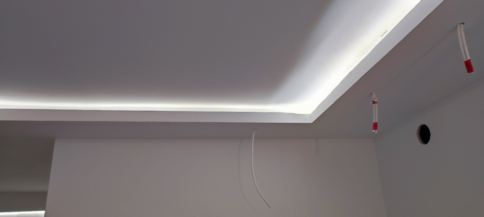 Nowoczesny sufit podwieszany z oświetleniem LED w narożniku, widoczne wystające przewody elektryczne i otwór w ścianie