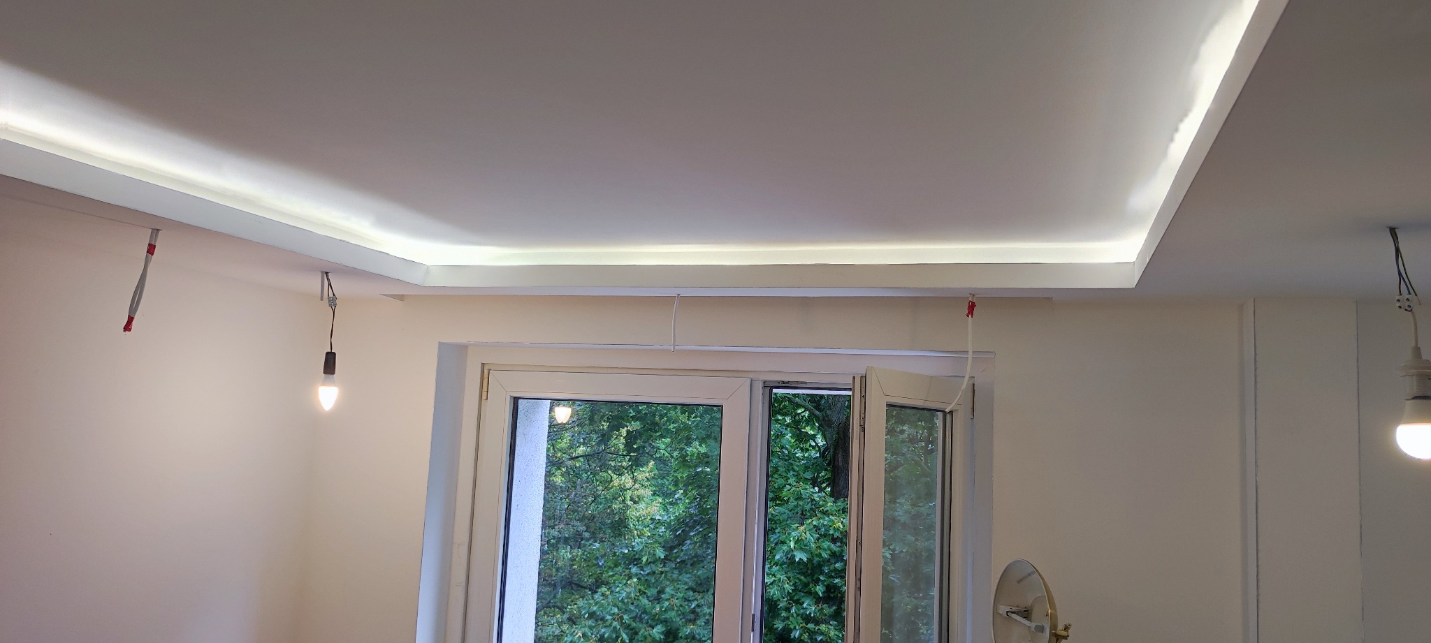 Biały sufit podwieszany z oświetleniem LED w narożnikach, widoczne okno z zielenią za szybą i wiszące żarówki na kablach.