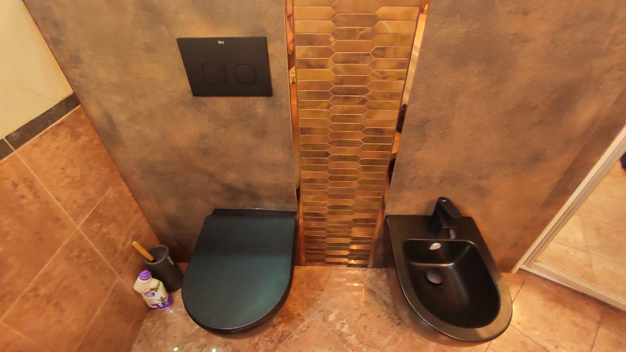 Wykończona łazienka z ciemną armaturą: wisząca toaleta z deską w kolorze grafitowym, bidet z czarną baterią, ściana wykończona płytami imitującymi beton oraz dekoracyjna wstawka z mozaiki...