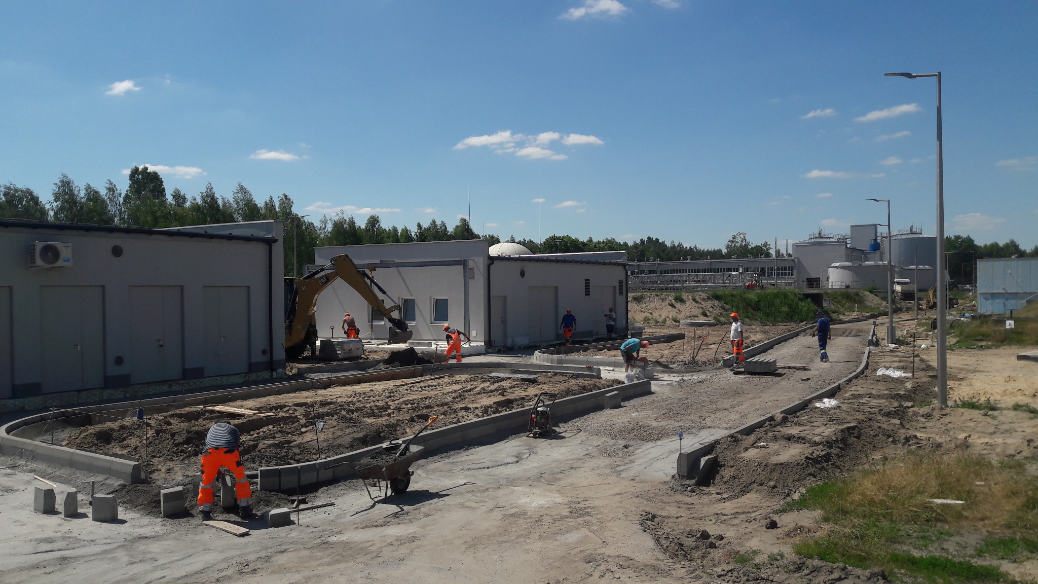 Budowa drogi dojazdowej na terenie przemysłowym, pracownicy w pomarańczowych kombinezonach układają obrzeża betonowe, w tle budynki i zbiorniki, na placu budowy stoi mała koparka.