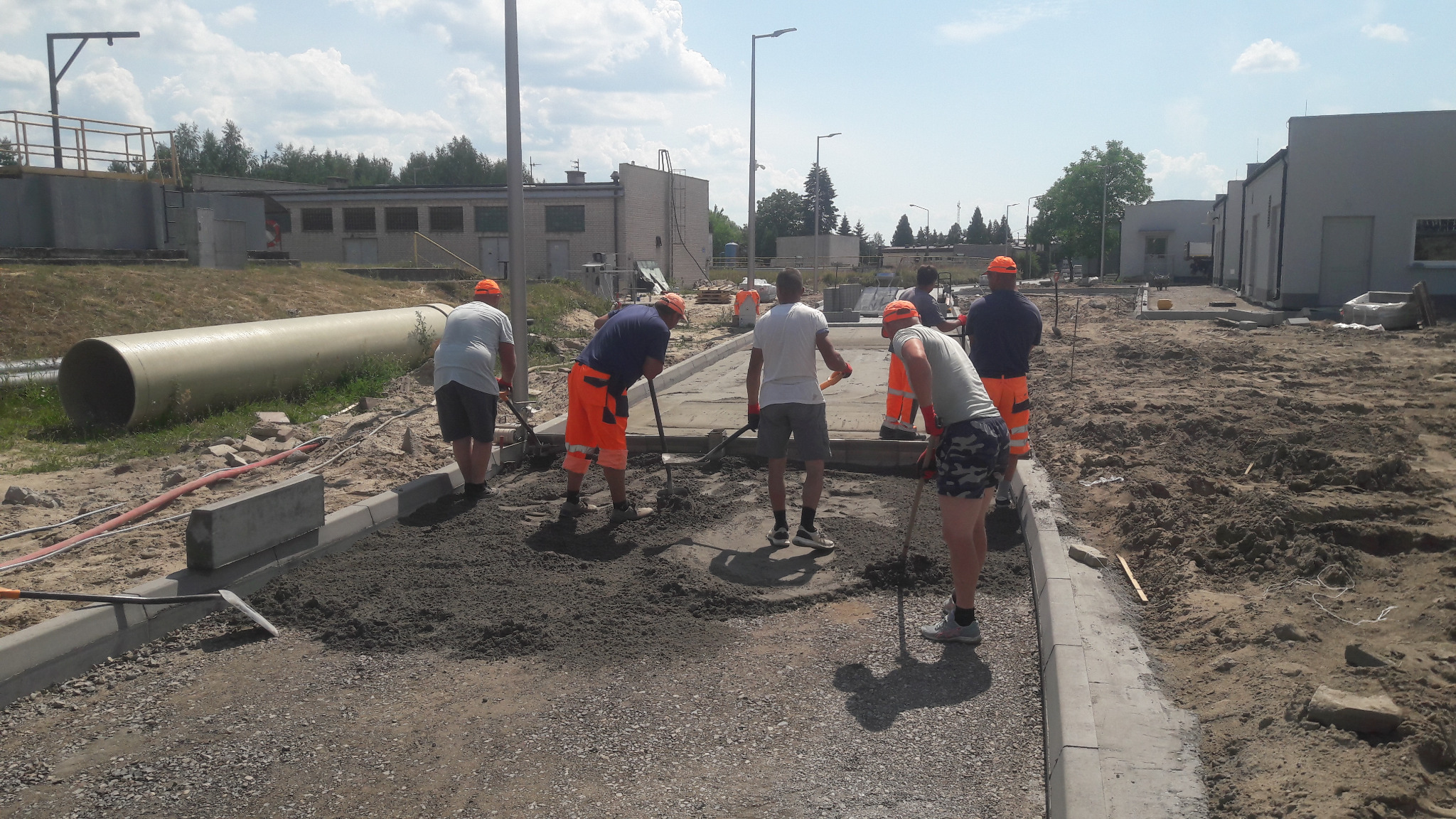 Ekipa robotników w pomarańczowych kamizelkach układa podłoże pod kostkę brukową na placu budowy w słoneczny dzień. Widoczne krawężniki, piasek i duża rura w tle.