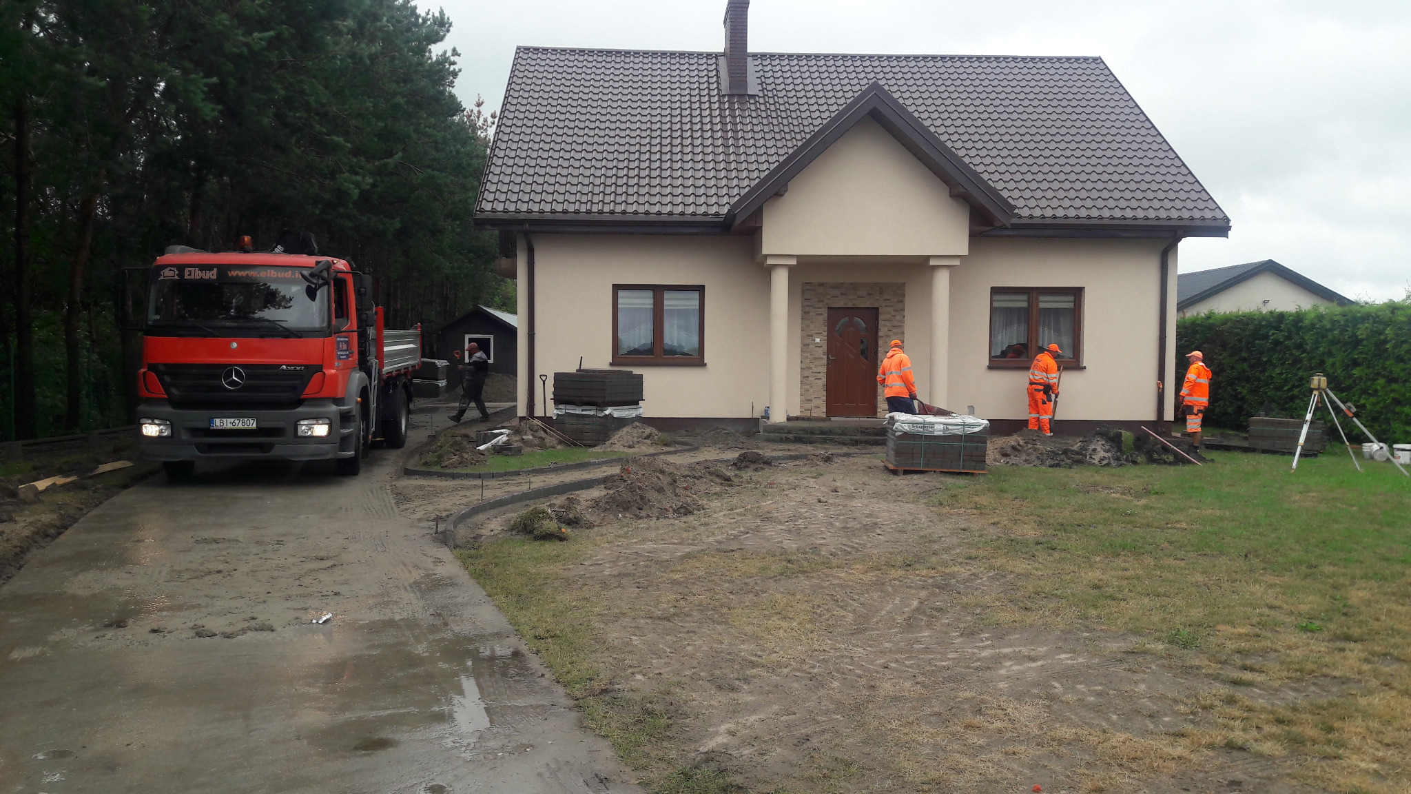 Prace brukarskie przed domem jednorodzinnym: ekipa w pomarańczowych kombinezonach układa obrzeża i przygotowuje podłoże pod kostkę, widoczna ciężarówka dostarczająca materiał, poziomica laserowa...