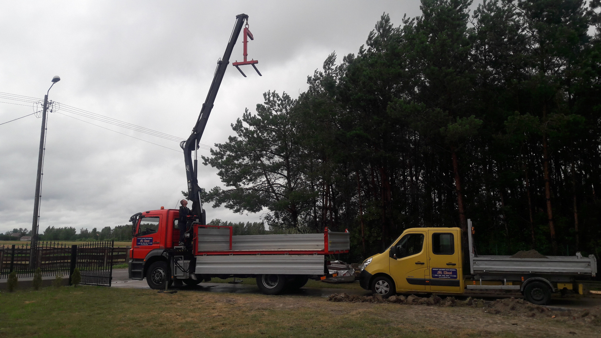Czerwona ciężarówka z dźwigiem i żółta furgonetka na trawiastym terenie, w tle las i pochmurne niebo.