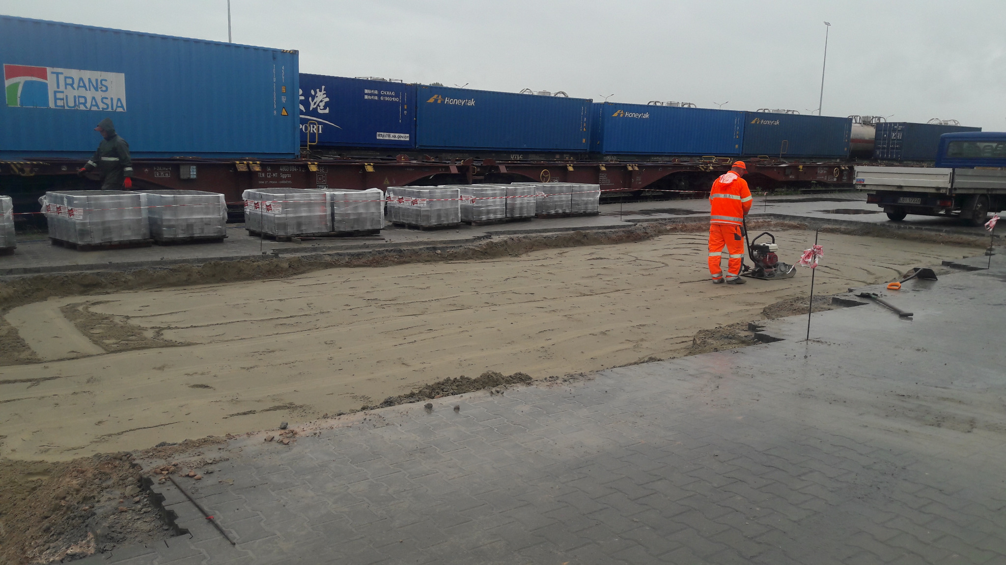 Pracownik w pomarańczowym kombinezonie zagęszcza podłoże pod kostkę brukową za pomocą zagęszczarki spalinowej; w tle pociąg towarowy z niebieskimi kontenerami.