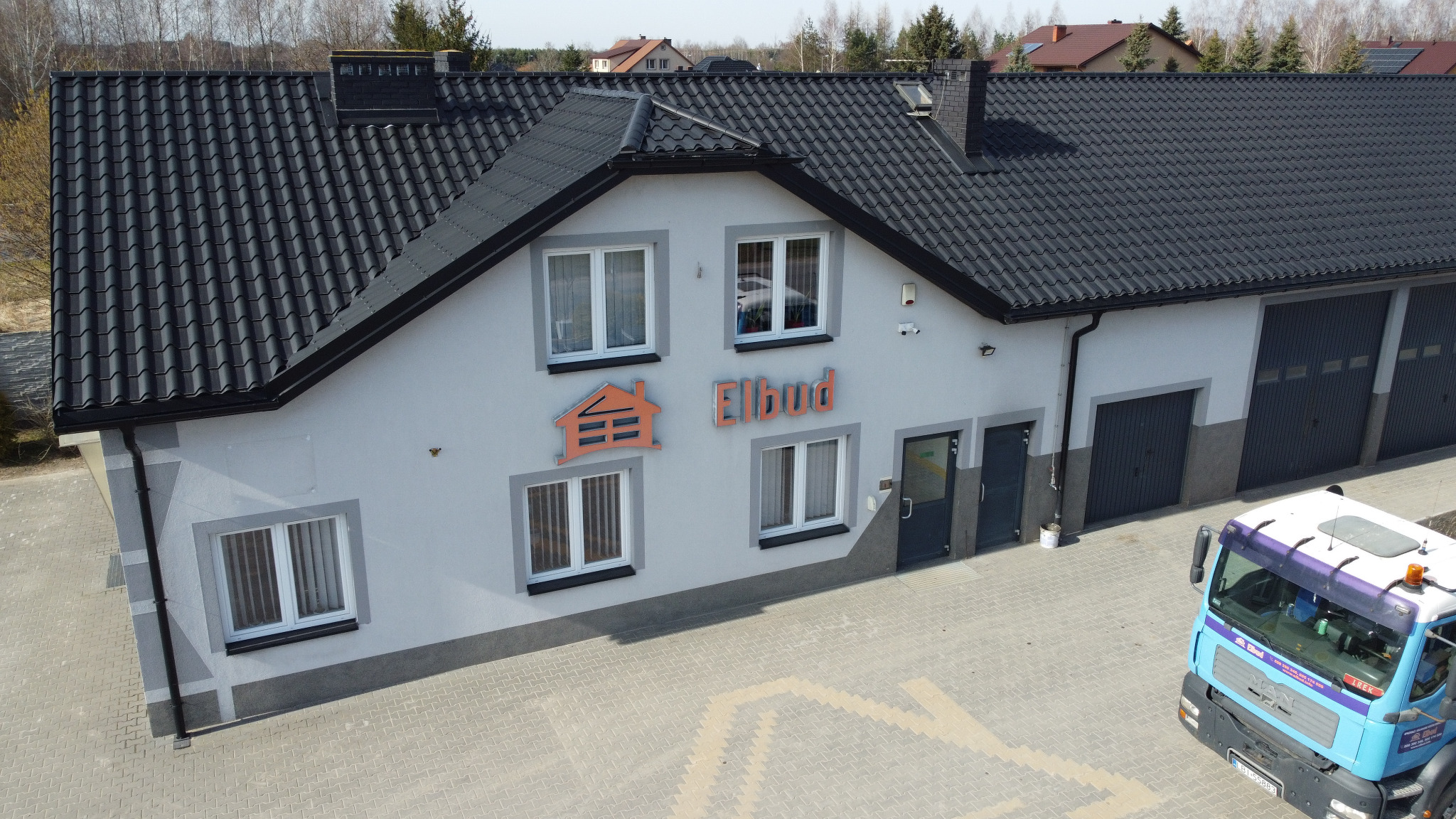 Budynek firmy z ciemną dachówką i logo 'Elbud' nad wejściem, z zaparkowaną ciężarówką na brukowanym podjeździe.