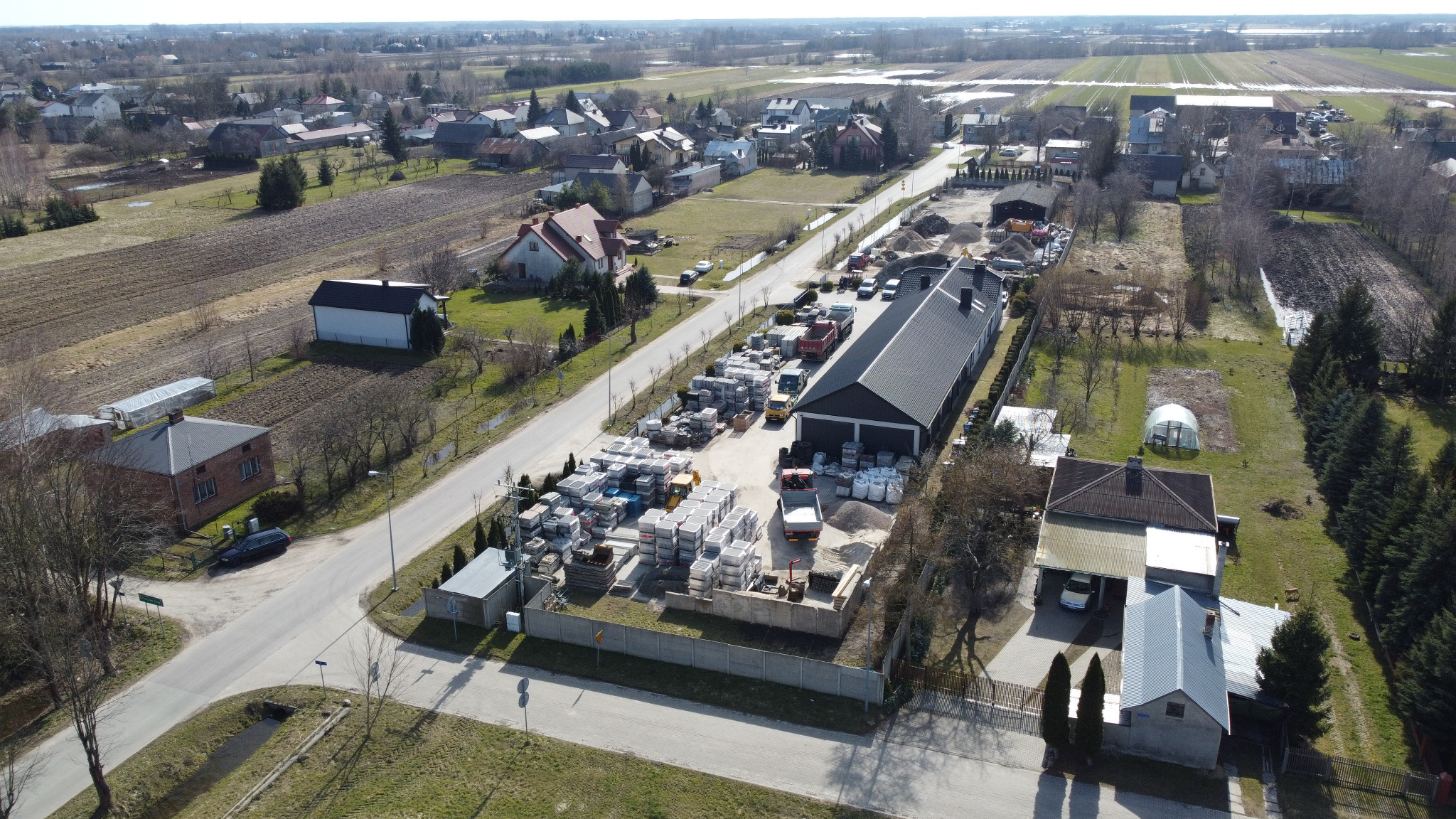Widok z lotu ptaka na skład kostki brukowej i materiałów budowlanych; palety z kostką, worki z kruszywem, ciężarówki i budynek magazynowy na obrzeżach wsi.