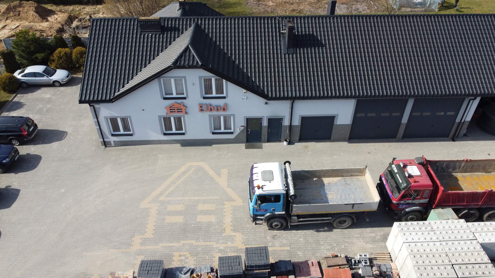 Widok z góry na budynek firmy Elbud z szarym dachem, zaparkowane samochody, dwa samochody ciężarowe z otwartymi skrzyniami ładunkowymi i ułożoną kostkę brukową z wzorem domu na placu.