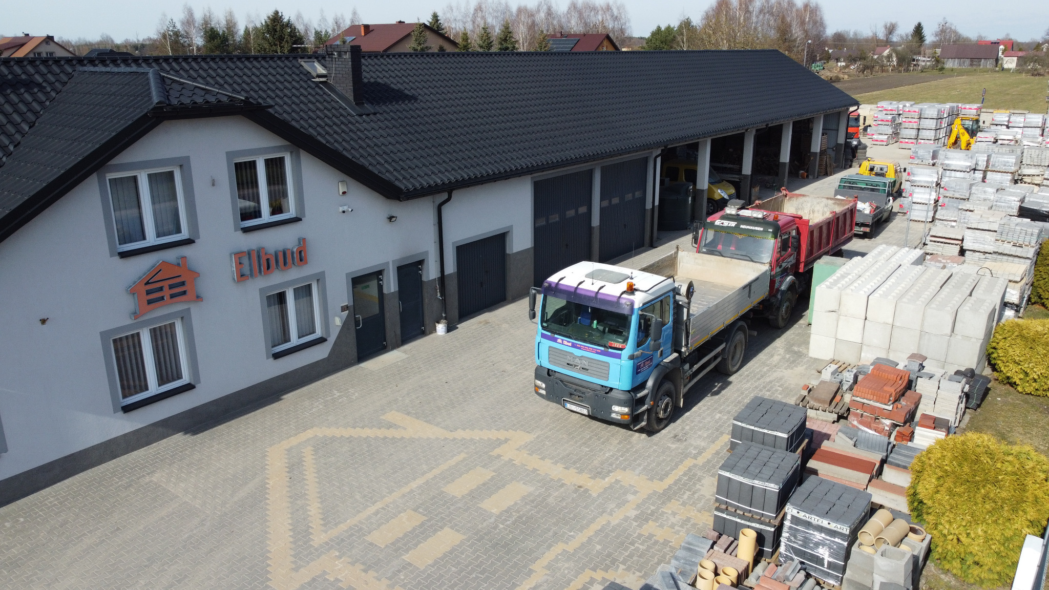 Widok z góry na plac firmy budowlanej Elbud z zaparkowanymi samochodami ciężarowymi, stertami kostki brukowej i budynkiem biurowym z logo firmy.