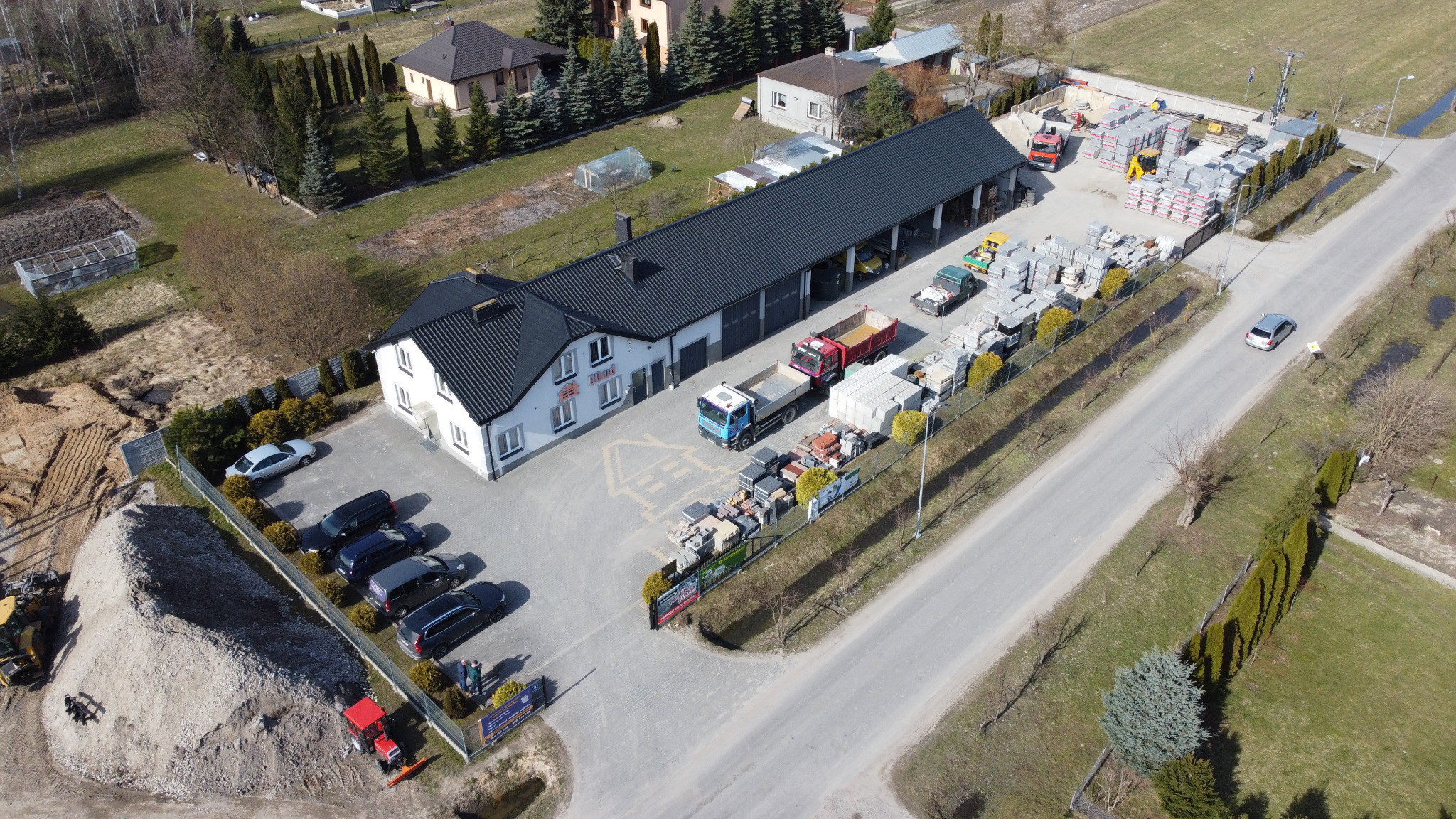 Szeroki kadr z lotu ptaka na firmę budowlaną z placem składowym kostki brukowej i ogrodzeń, zaparkowanymi samochodami osobowymi i ciężarowymi oraz stertą piasku z koparką w tle, widoczne budynki...