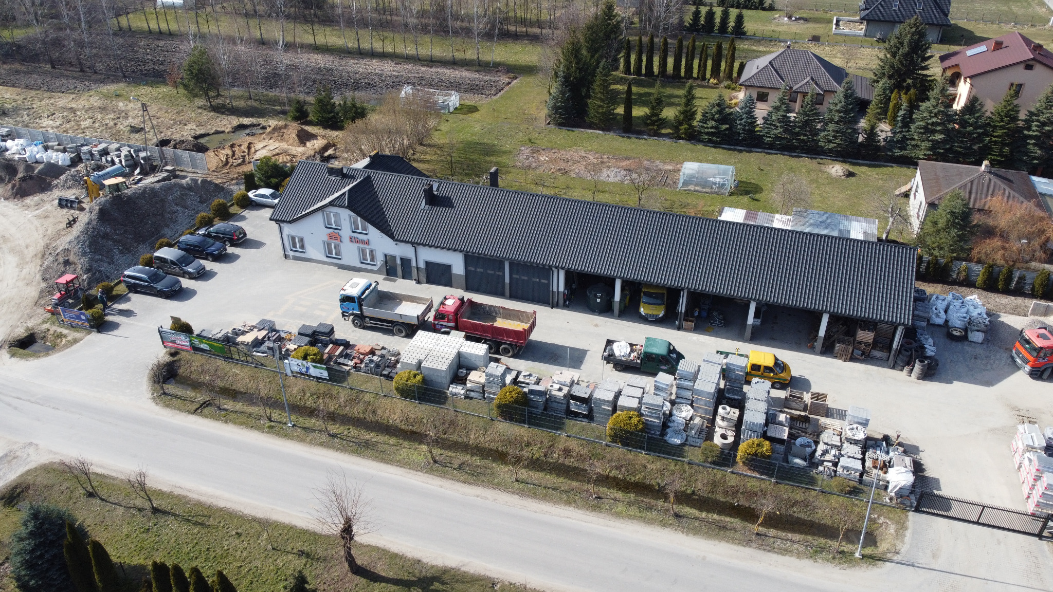 Widok z lotu ptaka na siedzibę firmy budowlanej z zapleczem: sterty kostki brukowej, samochody dostawcze, budynek biurowy z logo firmy Elbud, hałda ziemi i zaparkowane samochody osobowe.