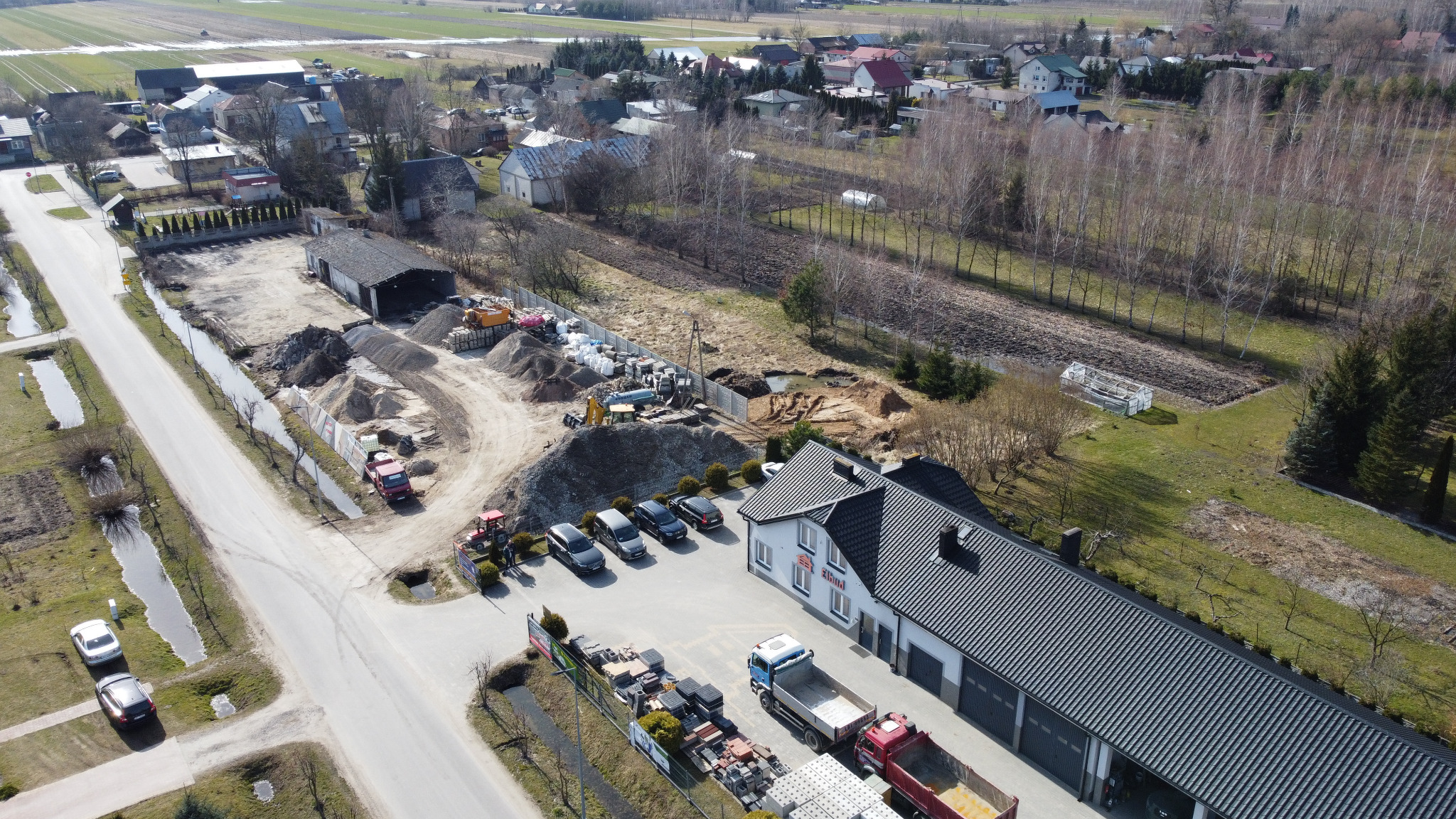 Widok z lotu ptaka na plac budowy z budynkiem firmy, zaparkowanymi samochodami, stertami gruzu, wykopem ziemnym, ciężarówkami i traktorem, w tle zabudowa wiejska i pola.