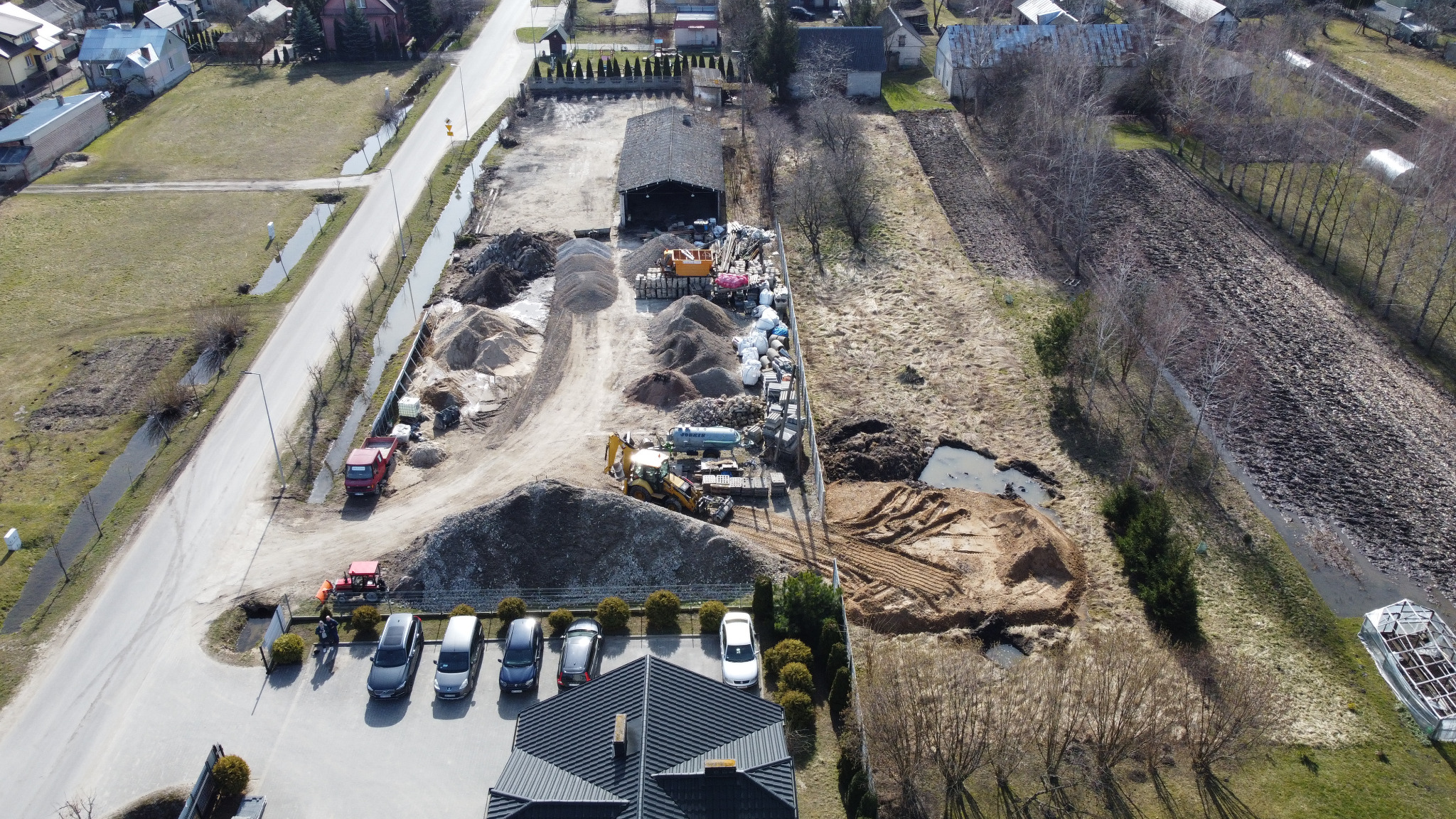 Widok z góry na plac budowy z hałdami piasku, żwiru, zaparkowanymi maszynami budowlanymi, wykopem, budynkiem gospodarczym i kilkoma samochodami osobowymi na parkingu.