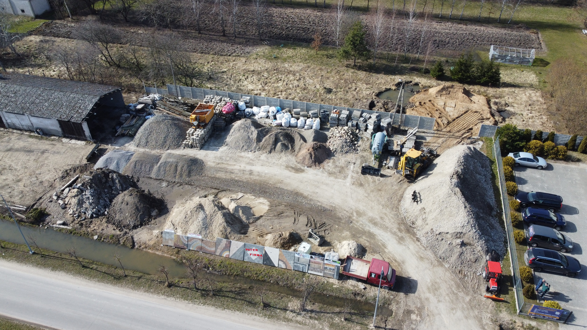 Szeroki kadr z lotu ptaka na plac budowy z maszynami, stertami materiałów budowlanych, kostką brukową, pojazdami i częściowo ogrodzonym terenem.