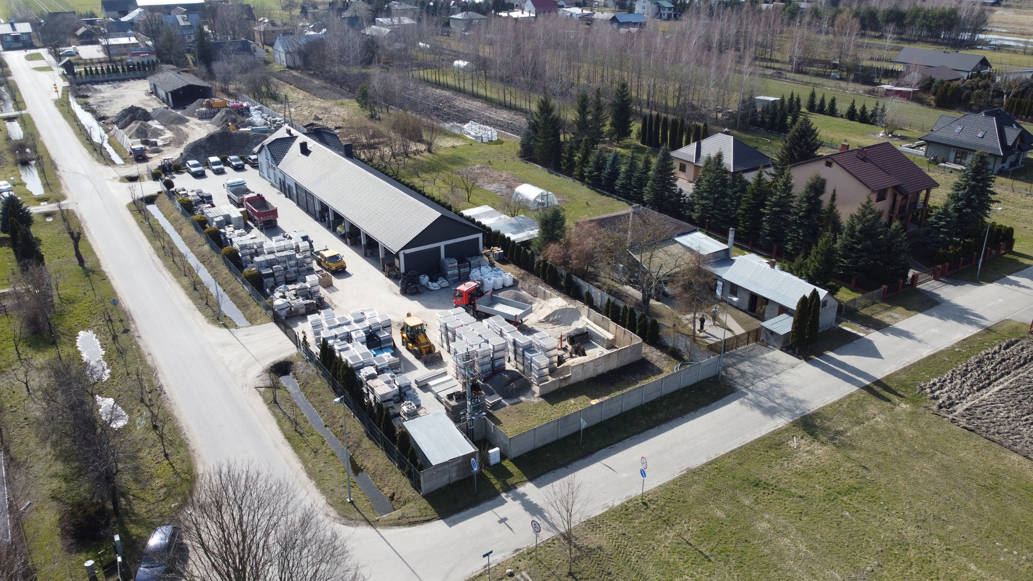 Widok z drona na skład kostki brukowej i materiałów budowlanych z zaparkowanymi pojazdami i budynkiem magazynowym, otoczony zielenią i zabudową jednorodzinną w tle.