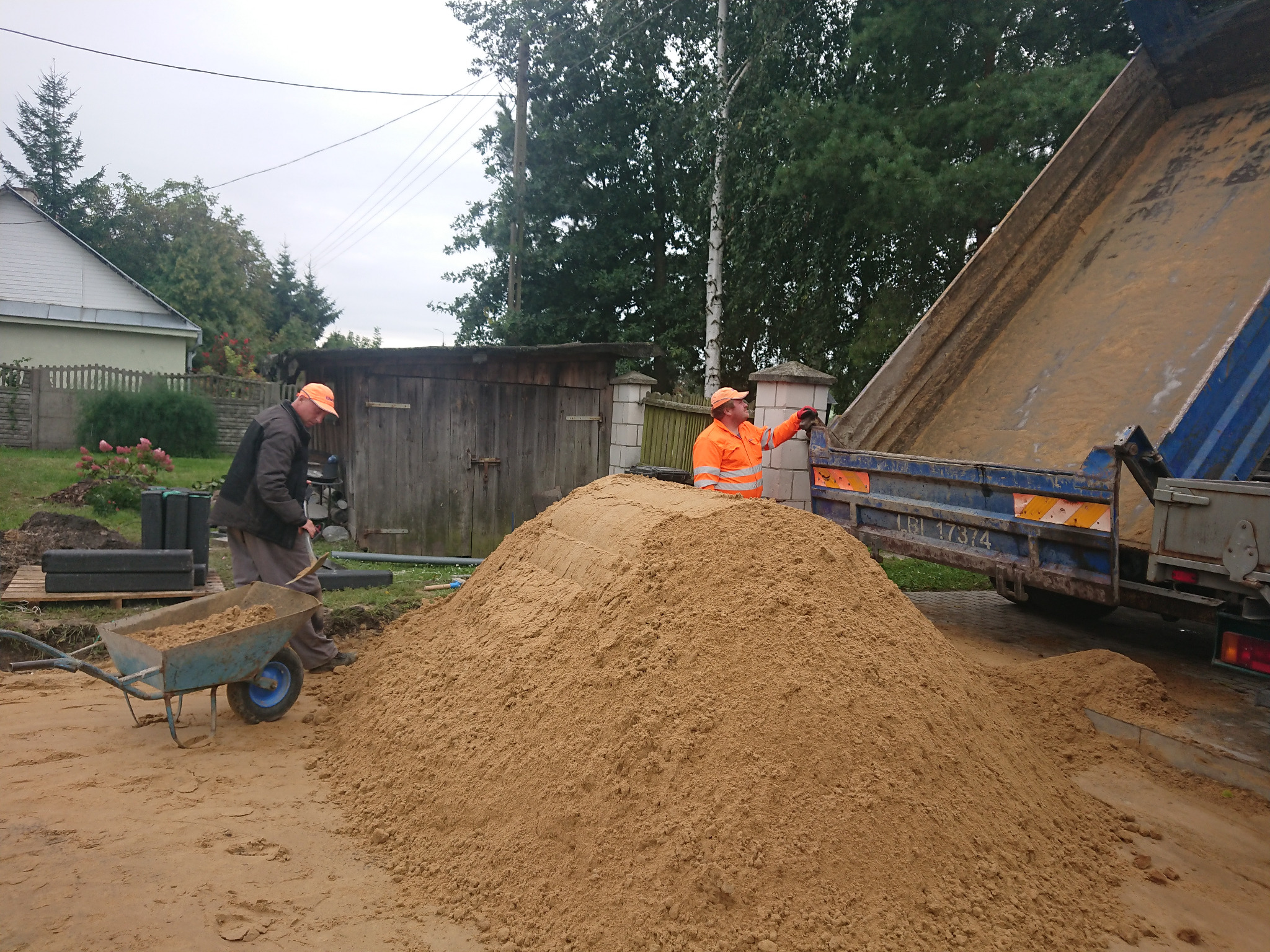Dwaj pracownicy w odblaskowych ubraniach pracują przy hałdzie piasku wysypanej z wywrotki na posesji z domem i drewnianym płotem w tle. Jeden z nich używa taczki.