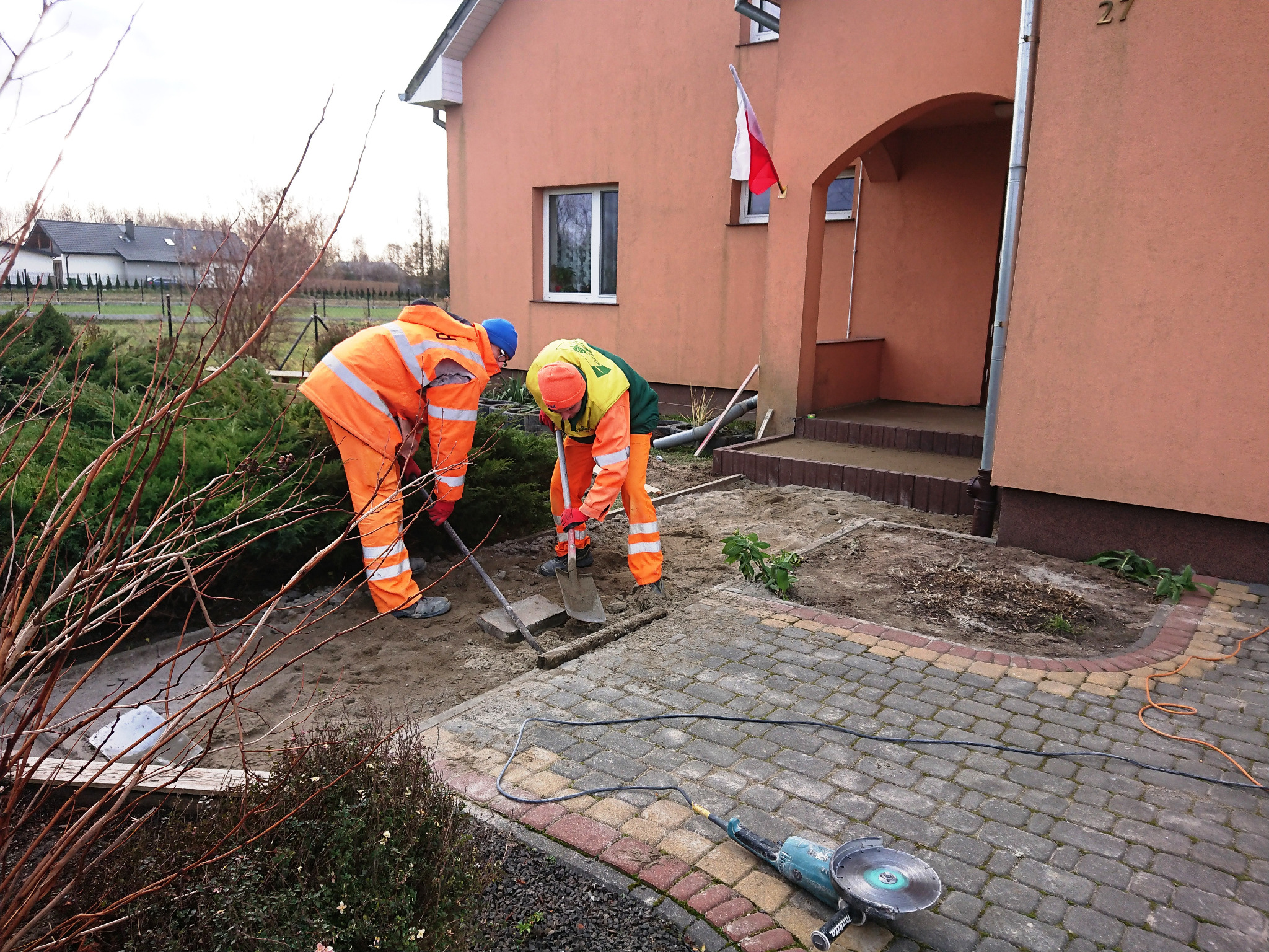 Dwóch pracowników w pomarańczowych kombinezonach przygotowuje podłoże pod kostkę brukową przed domem z numerem 27, używając łopat i szpadla. Na kostce leży szlifierka kątowa.