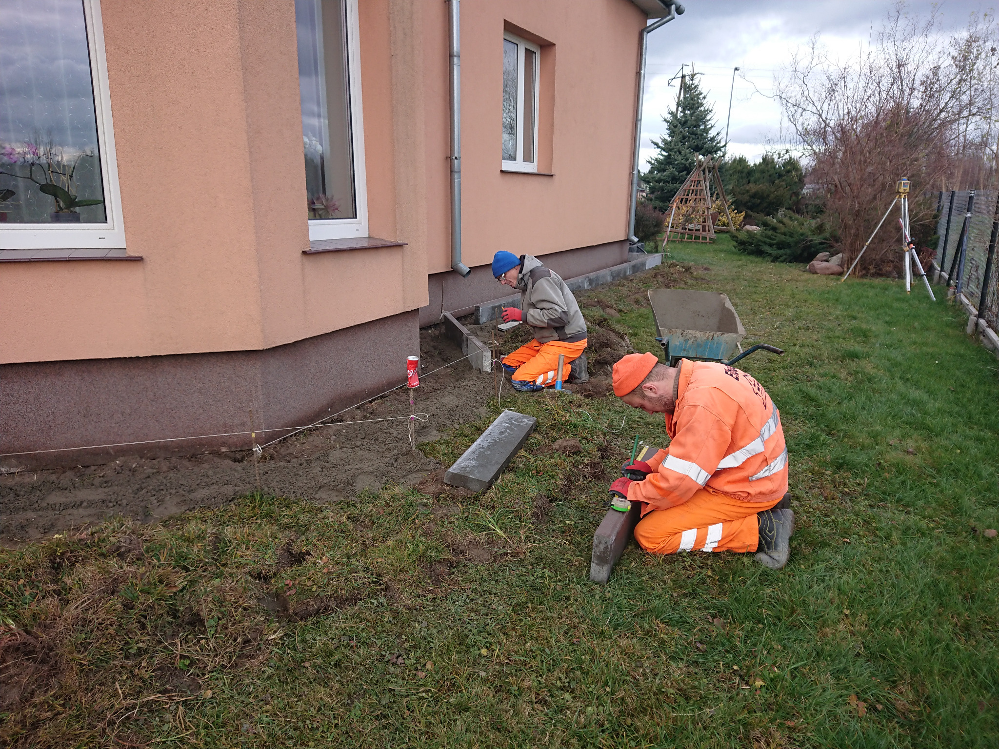 Dwóch pracowników w pomarańczowych kombinezonach układa obrzeża chodnika z szarej kostki przy fundamencie domu, używając poziomicy i sznurka wyznaczającego linię, z wózkiem budowlanym i niwelatorem...