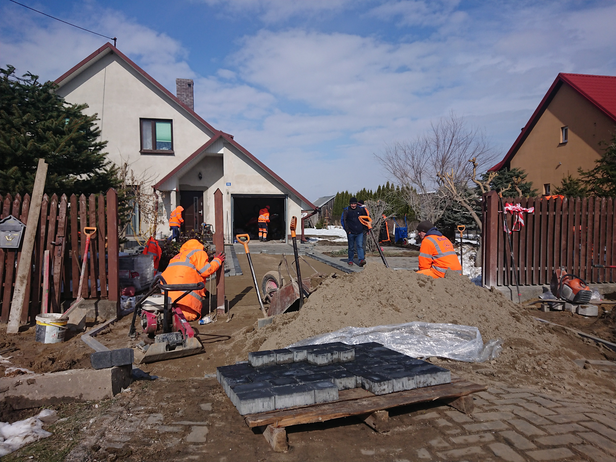 Układanie kostki brukowej na posesji: robotnicy w pomarańczowych kombinezonach pracują na terenie budowy, w tle domy i sterta piasku, przygotowane kostki brukowe na palecie.