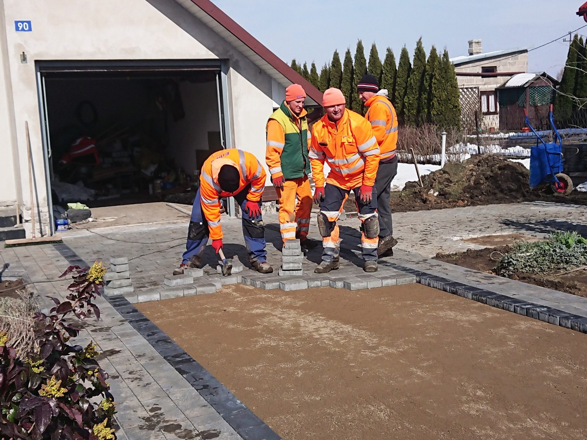 Pracownicy w odblaskowych ubraniach układają szarą kostkę brukową na podsypce piaskowej w Terespolu, widok z bliska na fragment nowej nawierzchni w trakcie tworzenia.